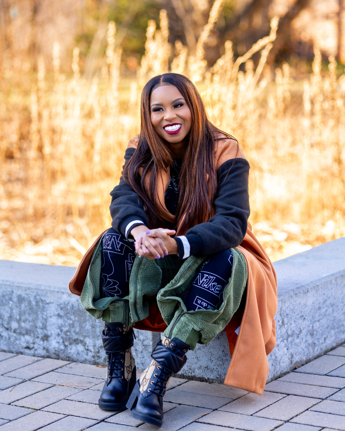 A smiling woman with long dark hair and bright lipstick squatting outside on a concrete bench, with a blurred background of autumn trees and fountain water. She is wearing a tan coat over a black hoodie, patterned pants, and Gucci and Gigi Hadid designer boots.