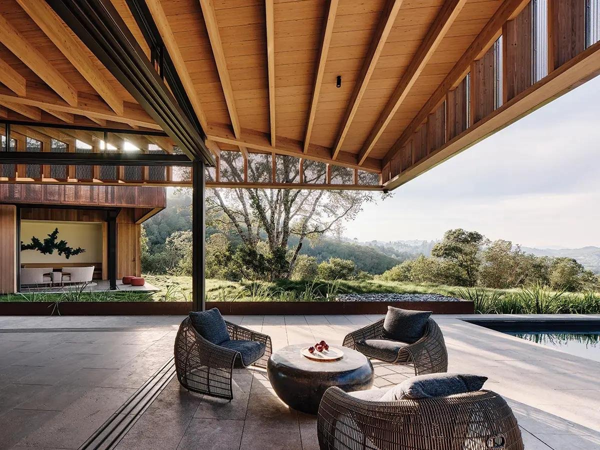 Modern outdoor patio with wicker chairs and a stone coffee table, overlooking a lush green landscape with trees and hills in the distance, under a wooden roof structure.