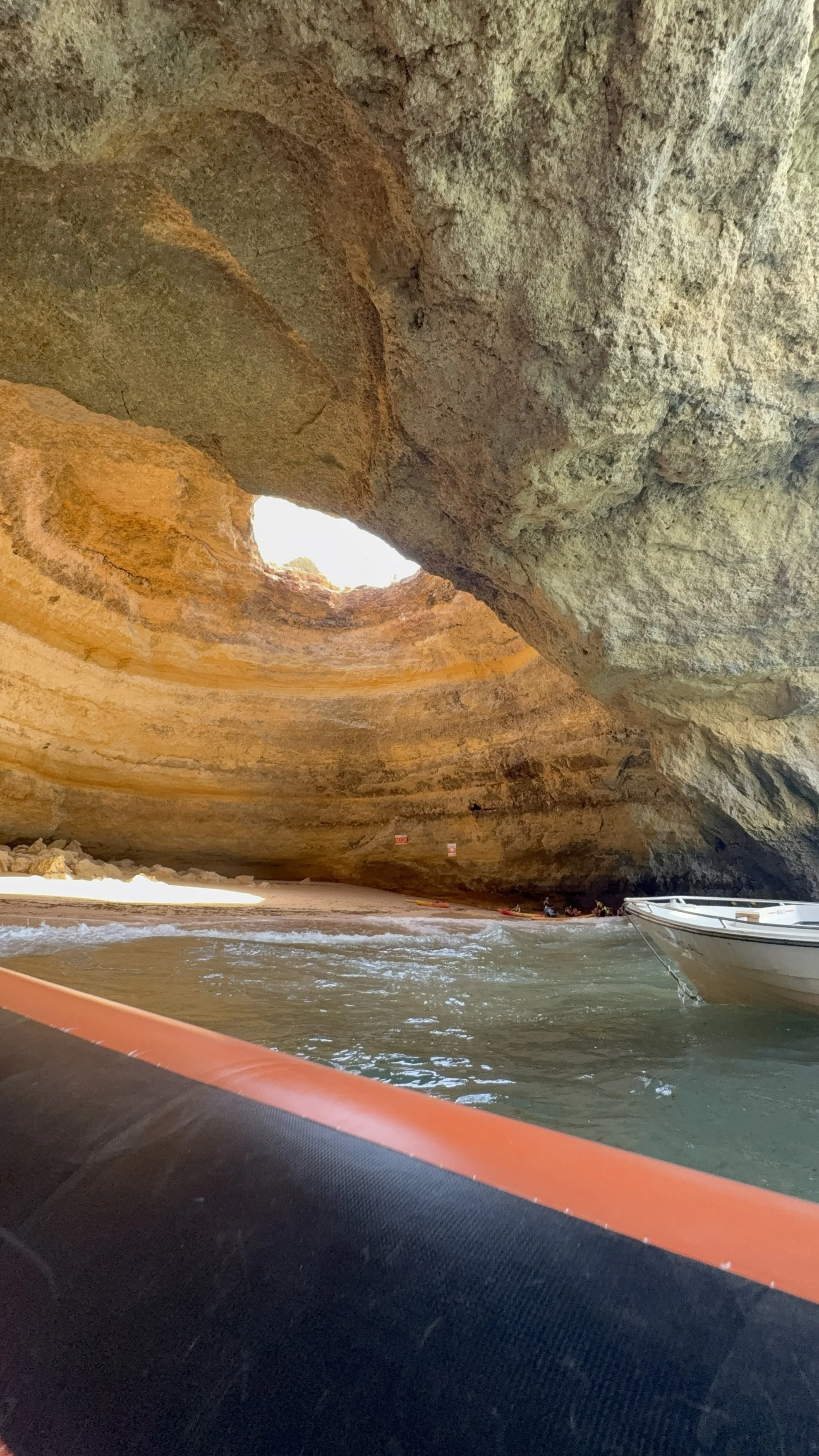 Inside a cave with a circular opening at the top, sunlight shining through, and water at the cave's base with a boat visible at the edge.
