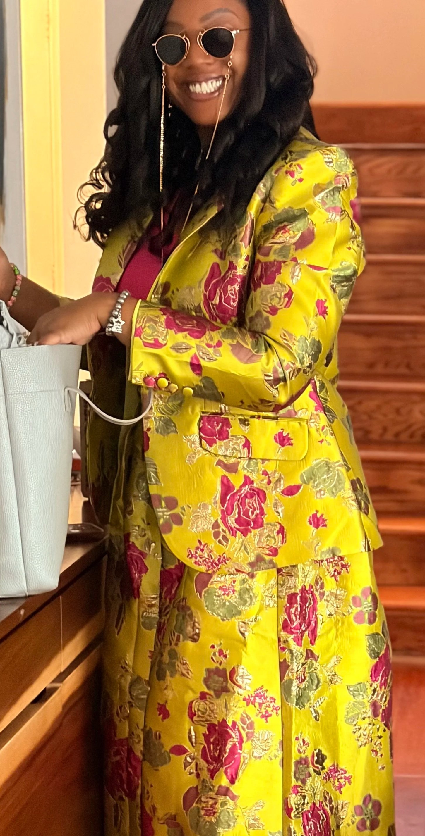 Woman in a bright yellow floral blazer and matching skirt, wearing sunglasses with gold chains, smiling, standing near a wooden staircase.