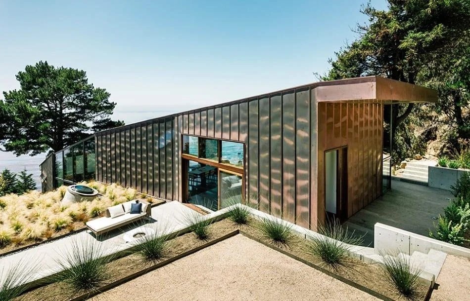 Modern house with copper exterior situated on a hillside with outdoor patio furniture and greenery, overlooking trees and the ocean.