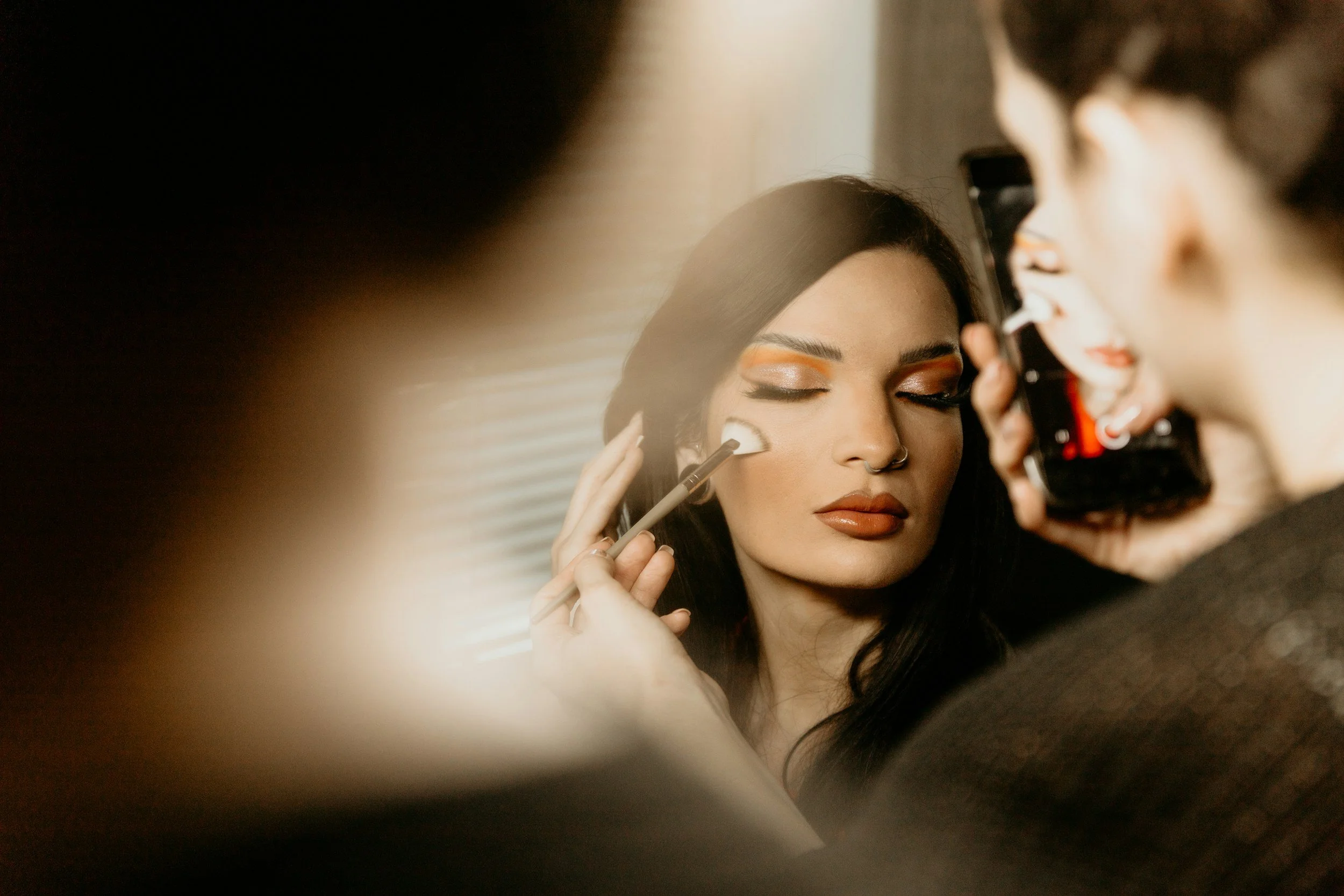 A woman with dark hair and makeup getting her eyeshadow applied by a makeup artist while looking into a mirror.