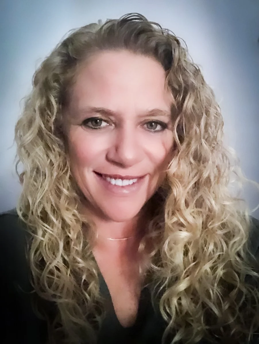 Close-up portrait of a woman with blonde curly hair, smiling, wearing a black top.