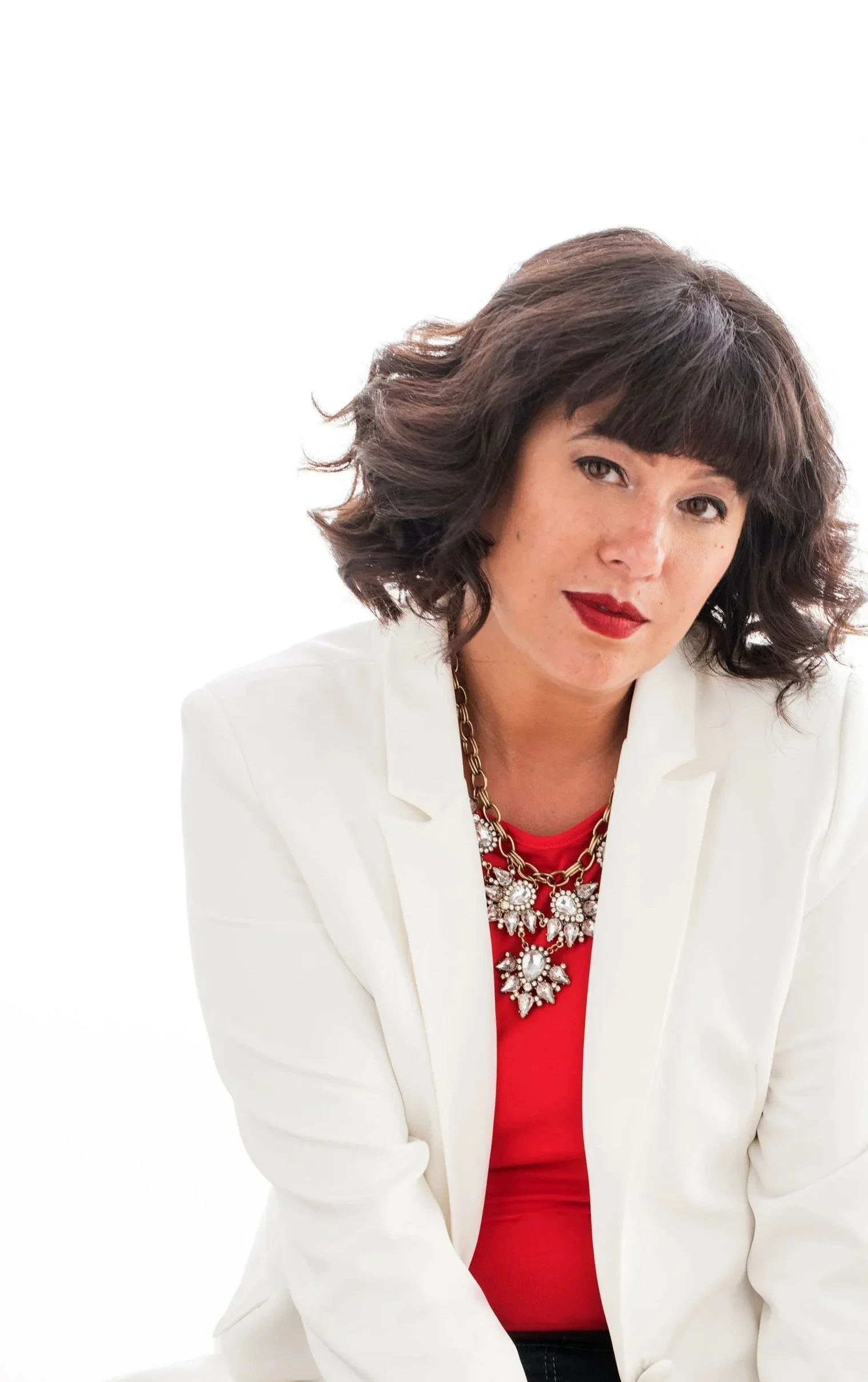 A woman with dark, wavy hair and red lipstick, wearing a white blazer over a red top and a statement necklace, posing against a white background.