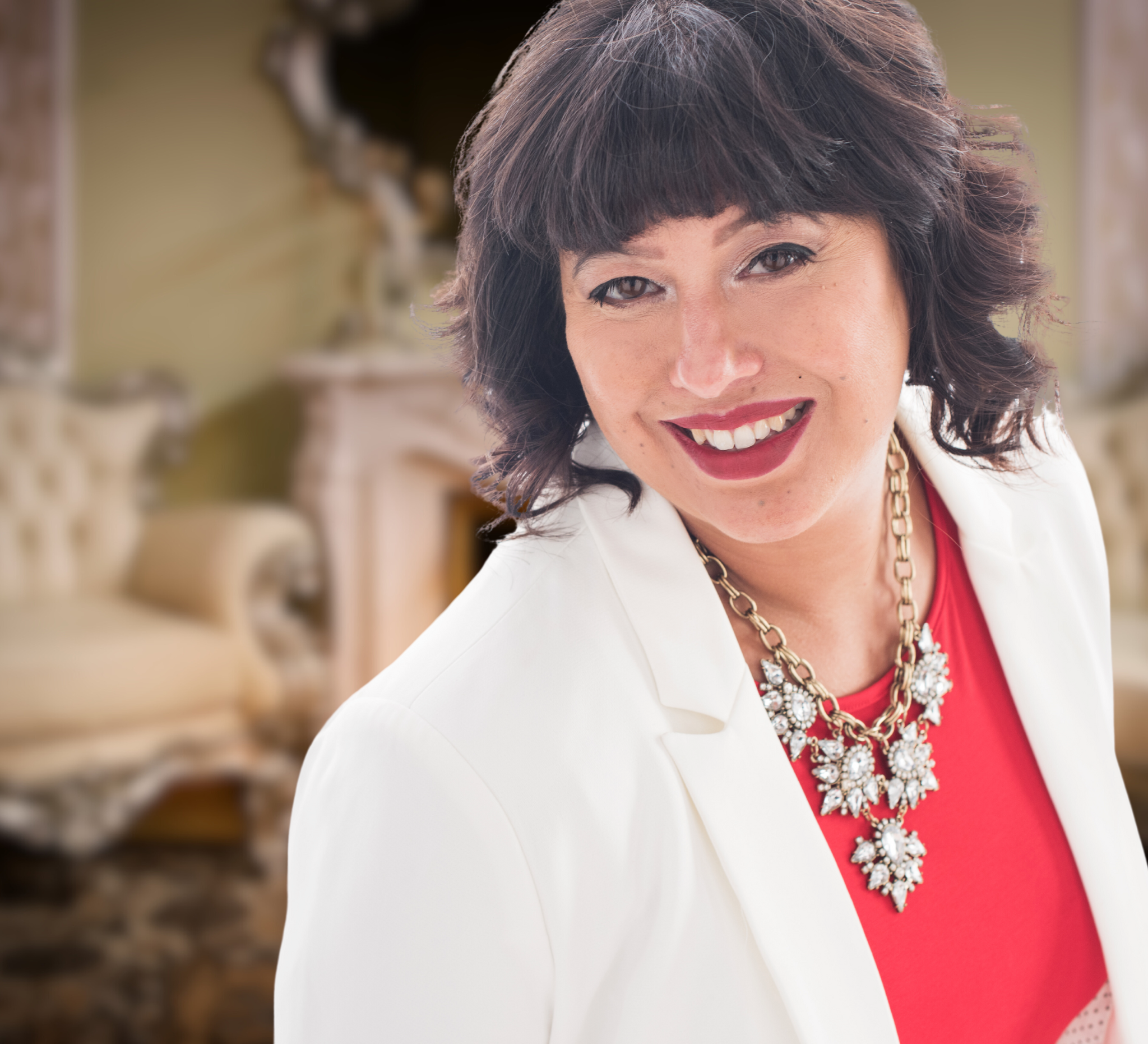 A woman with dark hair and a bright smile, wearing a white blazer, red top, and a jeweled necklace, standing in a luxurious room with a vintage sofa and ornate fireplace in the background.