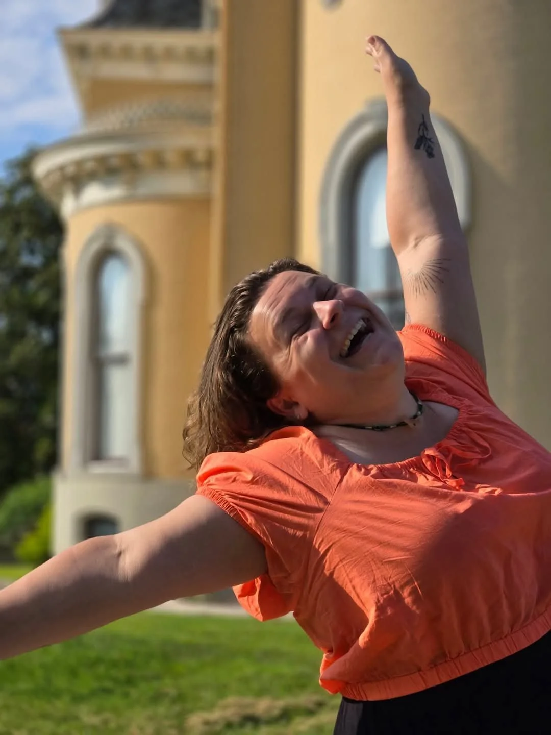 Woman wearing an orange top smiling with eyes closed and arms raised outdoors in front of a yellow building.