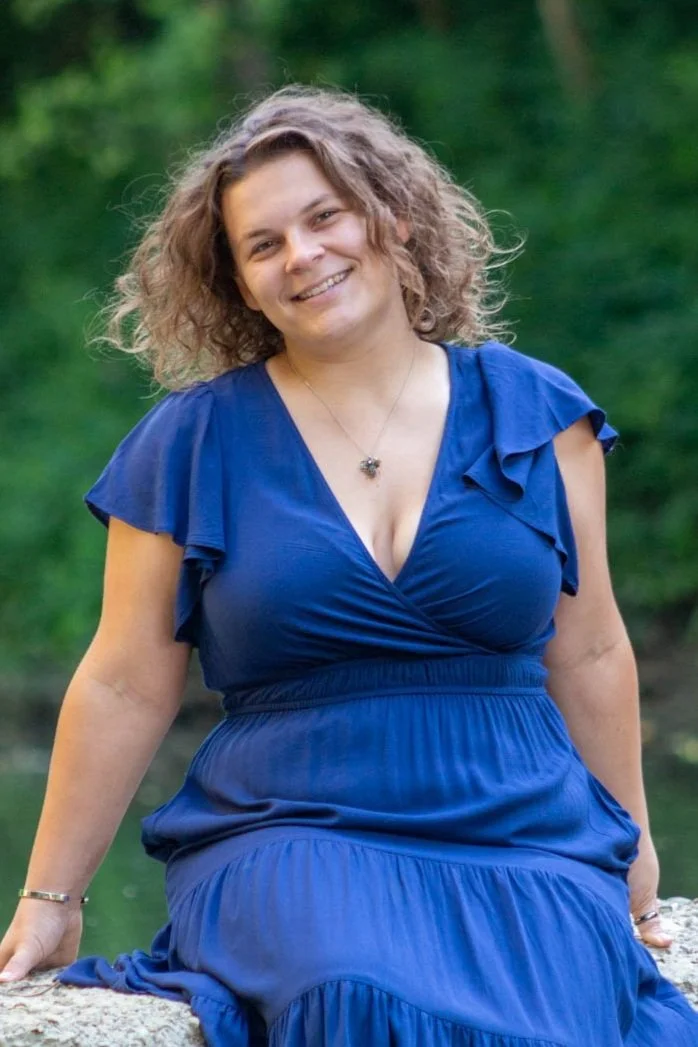 A smiling woman with curly hair wearing a blue dress outdoors near a body of water.