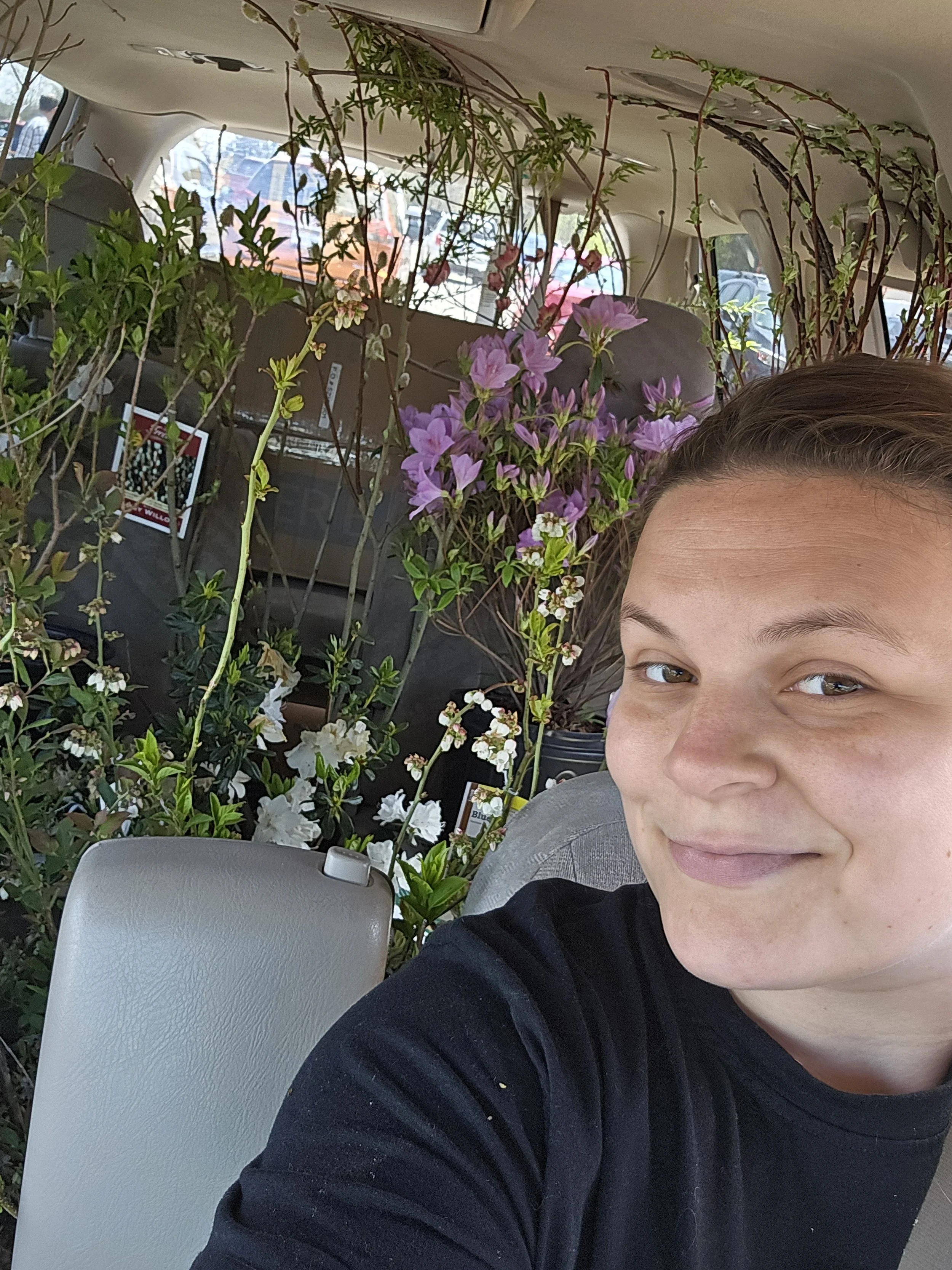 Partial view of a young person smiling in the foreground, with purple and white flowers and green plants behind them inside a vehicle.