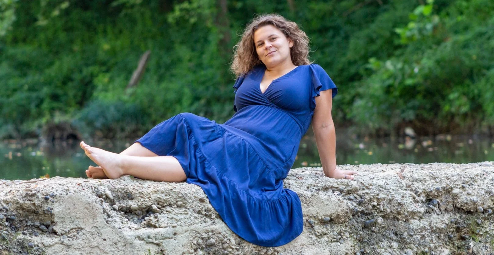 Woman in a blue dress sitting on a rocky ledge near water with greenery in the background.