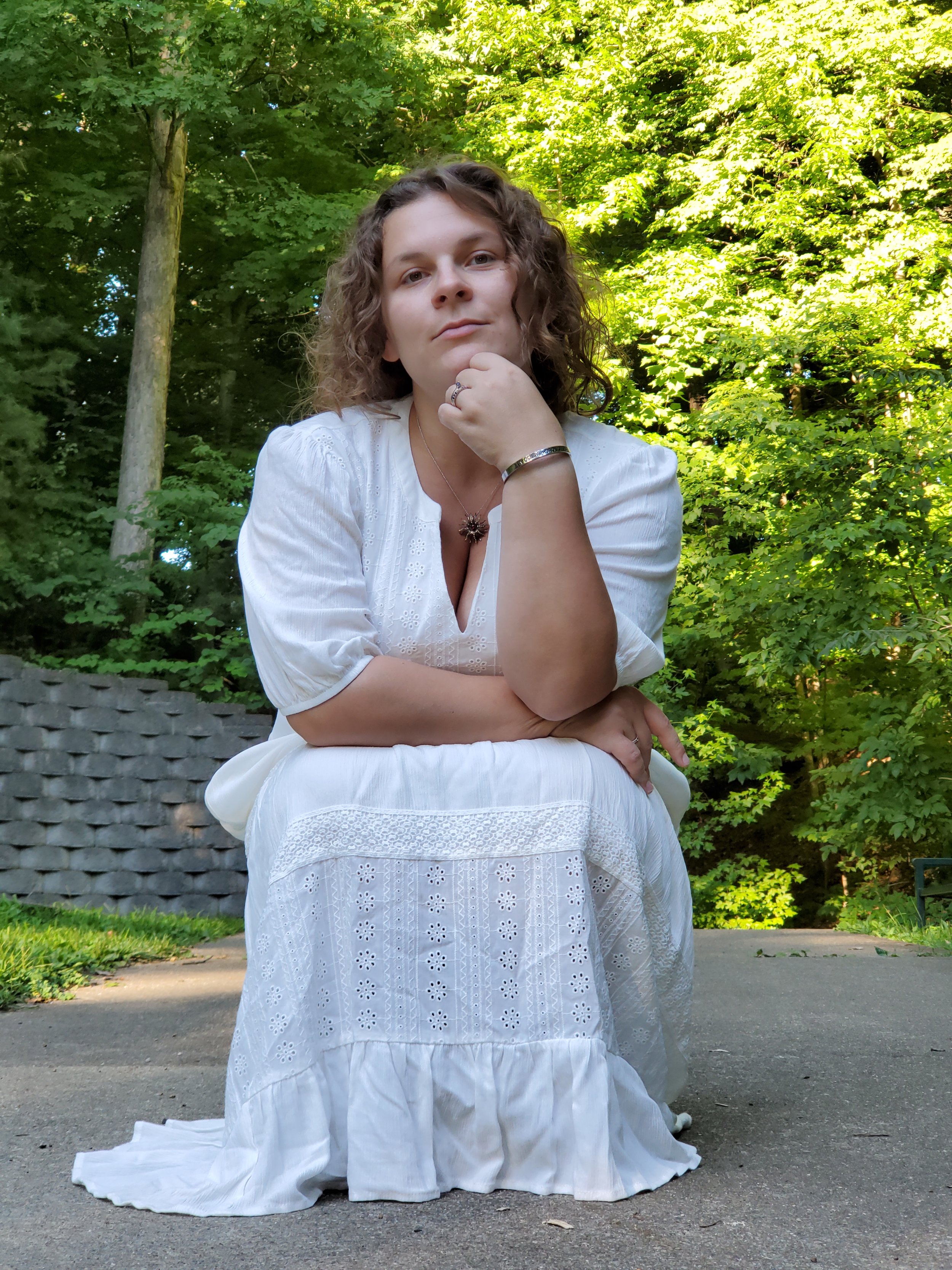 A woman with curly hair dressed in a white eyelet dress and wearing jewelry, sitting outdoors in front of a green leafy background, with her chin resting on her hand, looking directly at the camera.