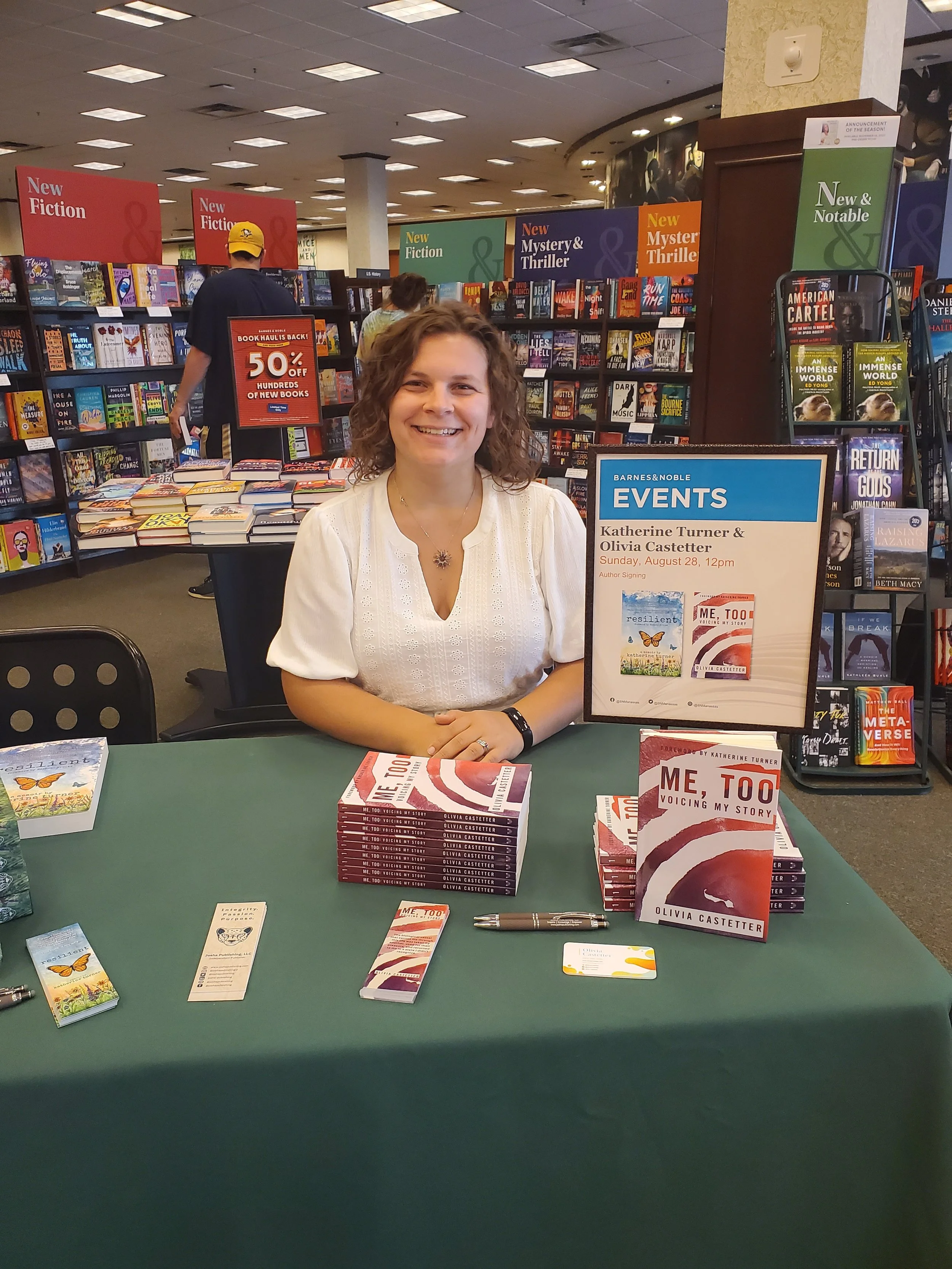 Author Olivia Castetter smiling at her book signing event in a bookstore, with copies of her book 'Me, Too' and promotional materials on the table.