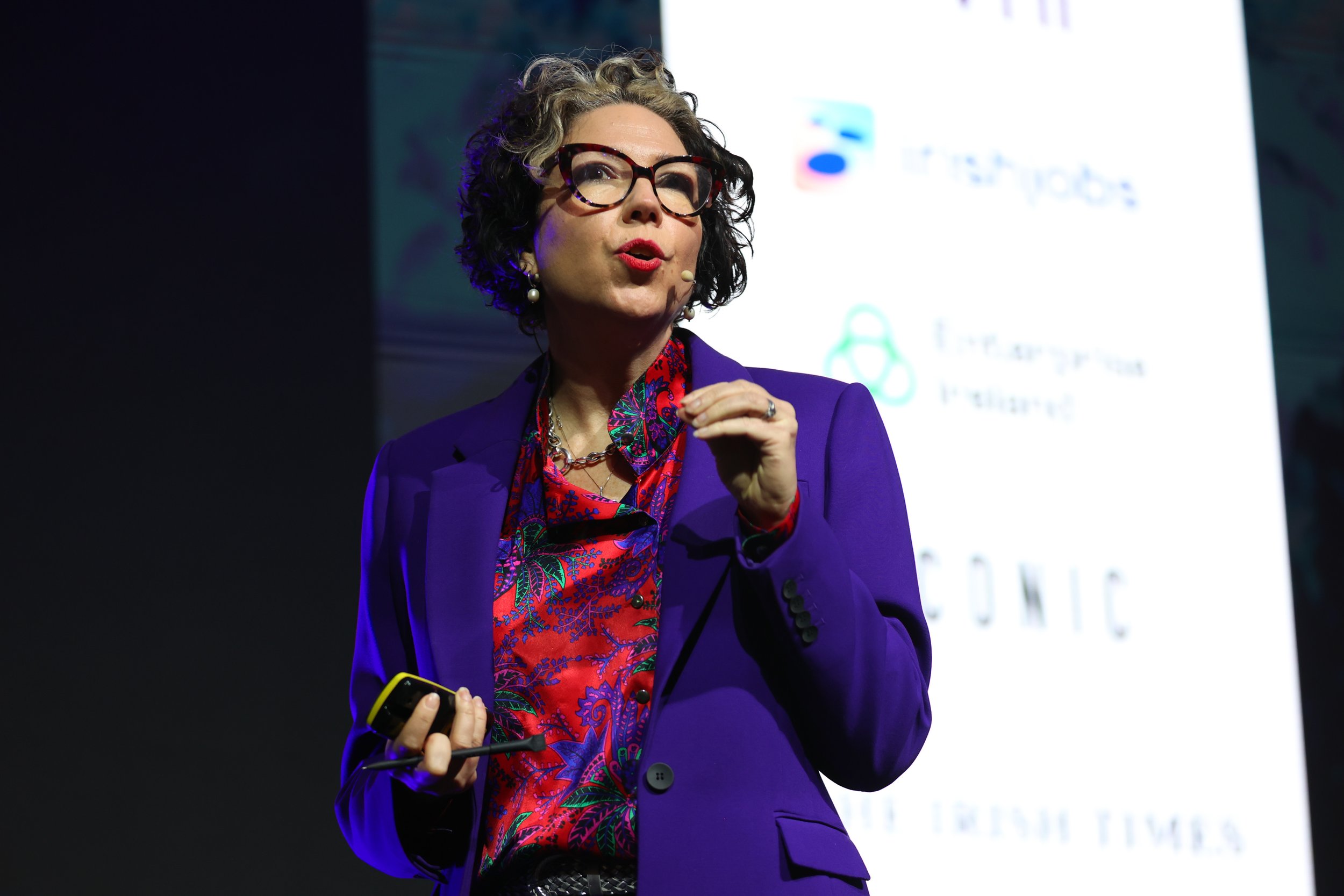 A woman wearing glasses and a purple blazer giving a presentation. She has curly hair and is holding a remote control.