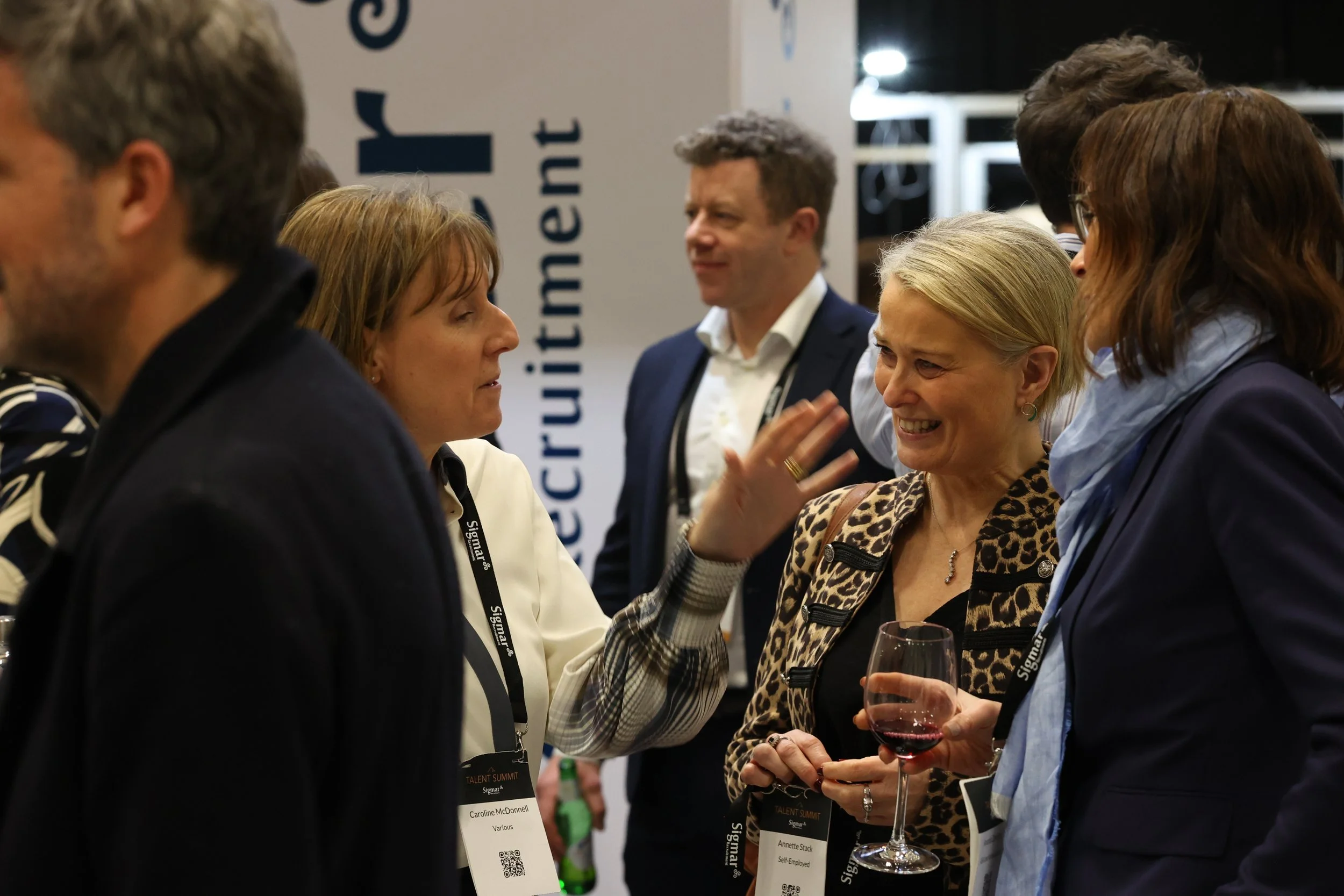 People are engaged in conversation at a professional networking event, several holding glasses of red wine, with name tags visible. There is a large sign in the background with partially visible text.