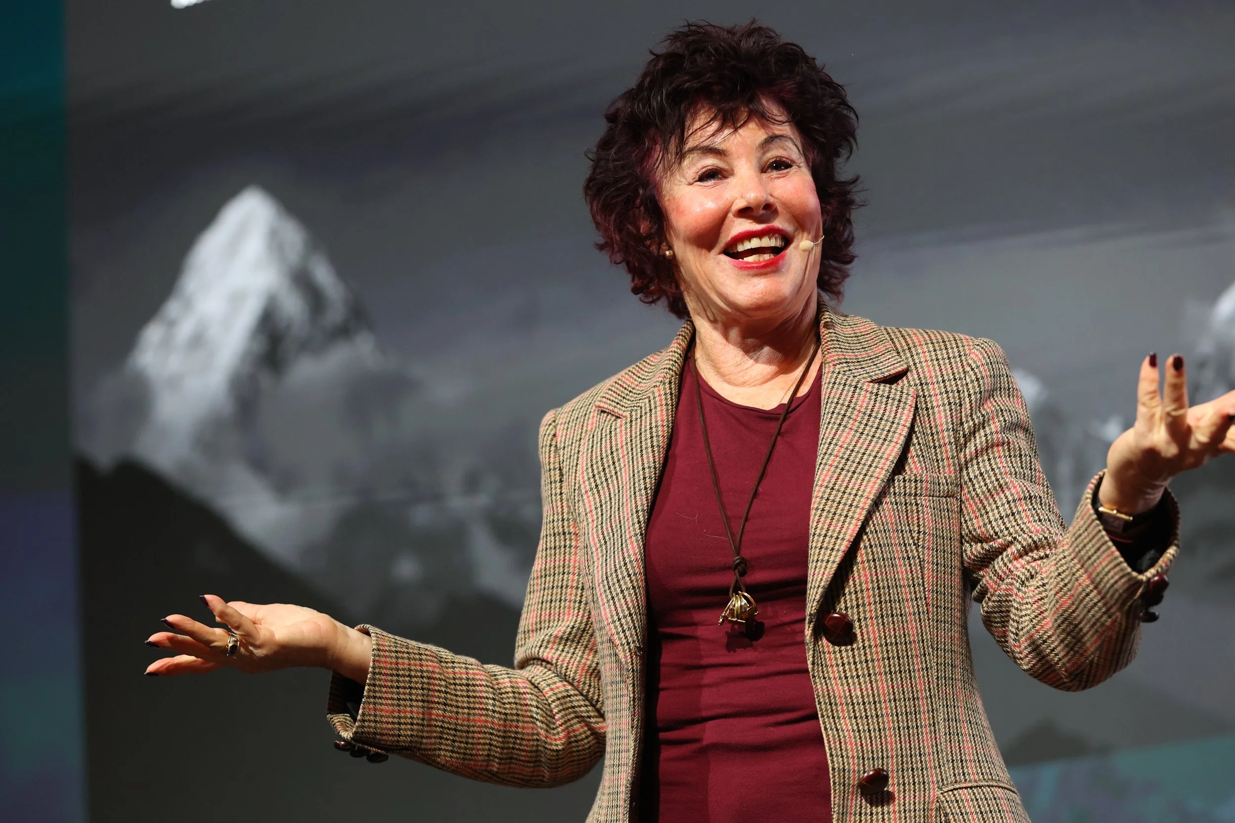 A woman with short curly brown hair, wearing a checkered blazer and red shirt, speaking at an event with mountains in the background.