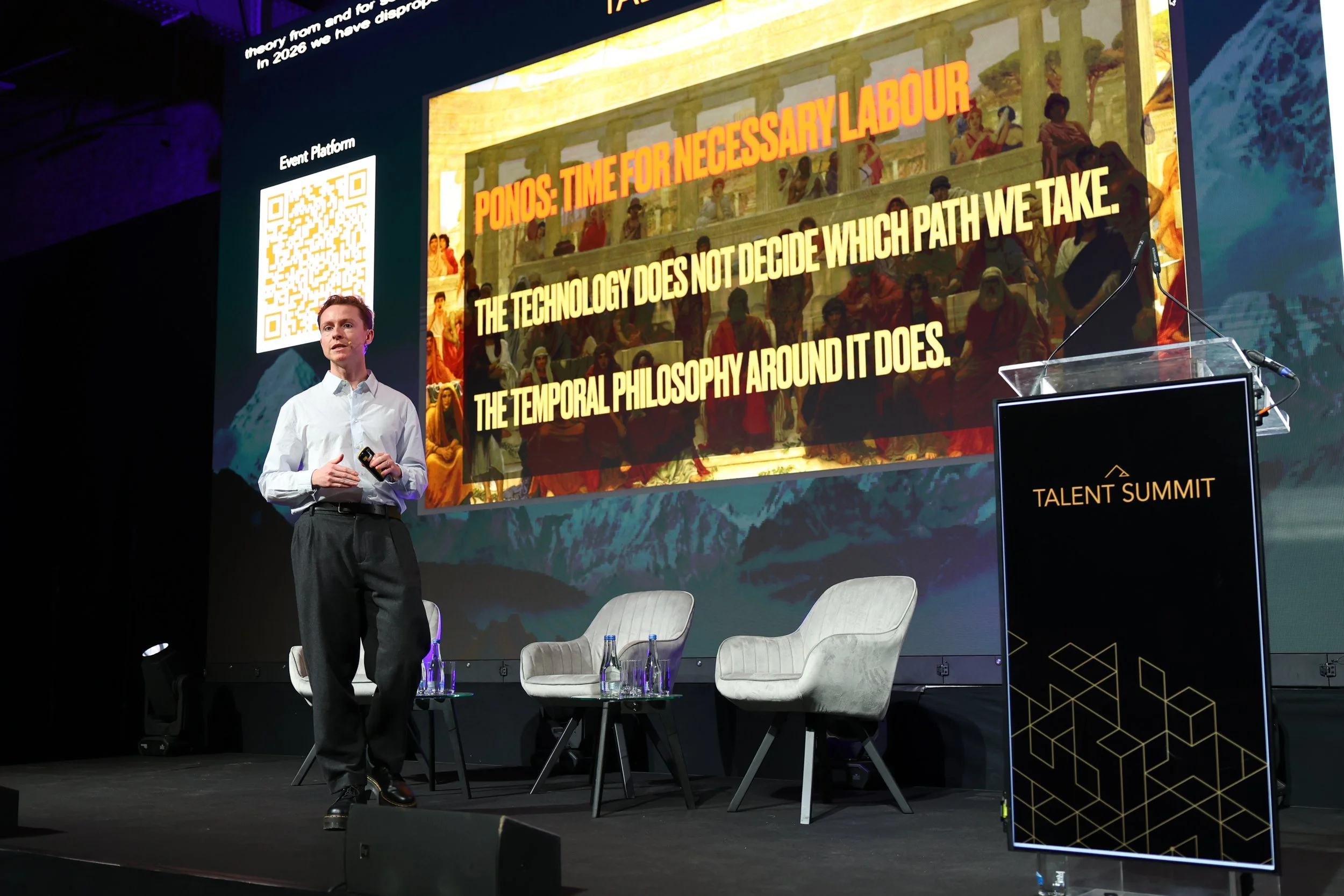 A man speaking at the Talent Summit, standing on stage with a large screen behind him displaying a slide with text and an artwork of people sitting in a forum. The slide reads: "PONOS: TIME FOR NECESSARY LABOUR. THE TECHNOLOGY DOES NOT DECIDE WHICH P
