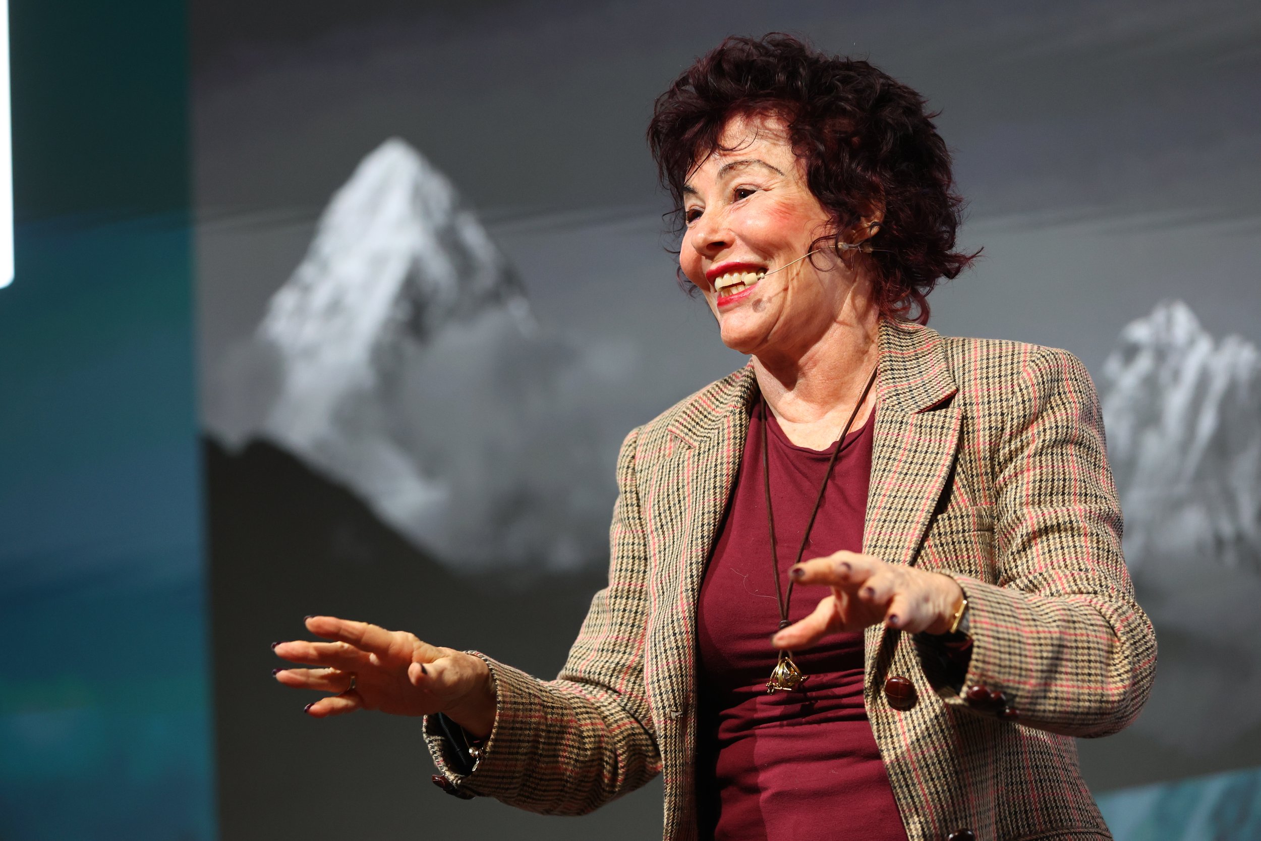 A woman with curly dark hair and a smile, wearing a plaid blazer and burgundy top, speaking at a conference with a mountain backdrop.