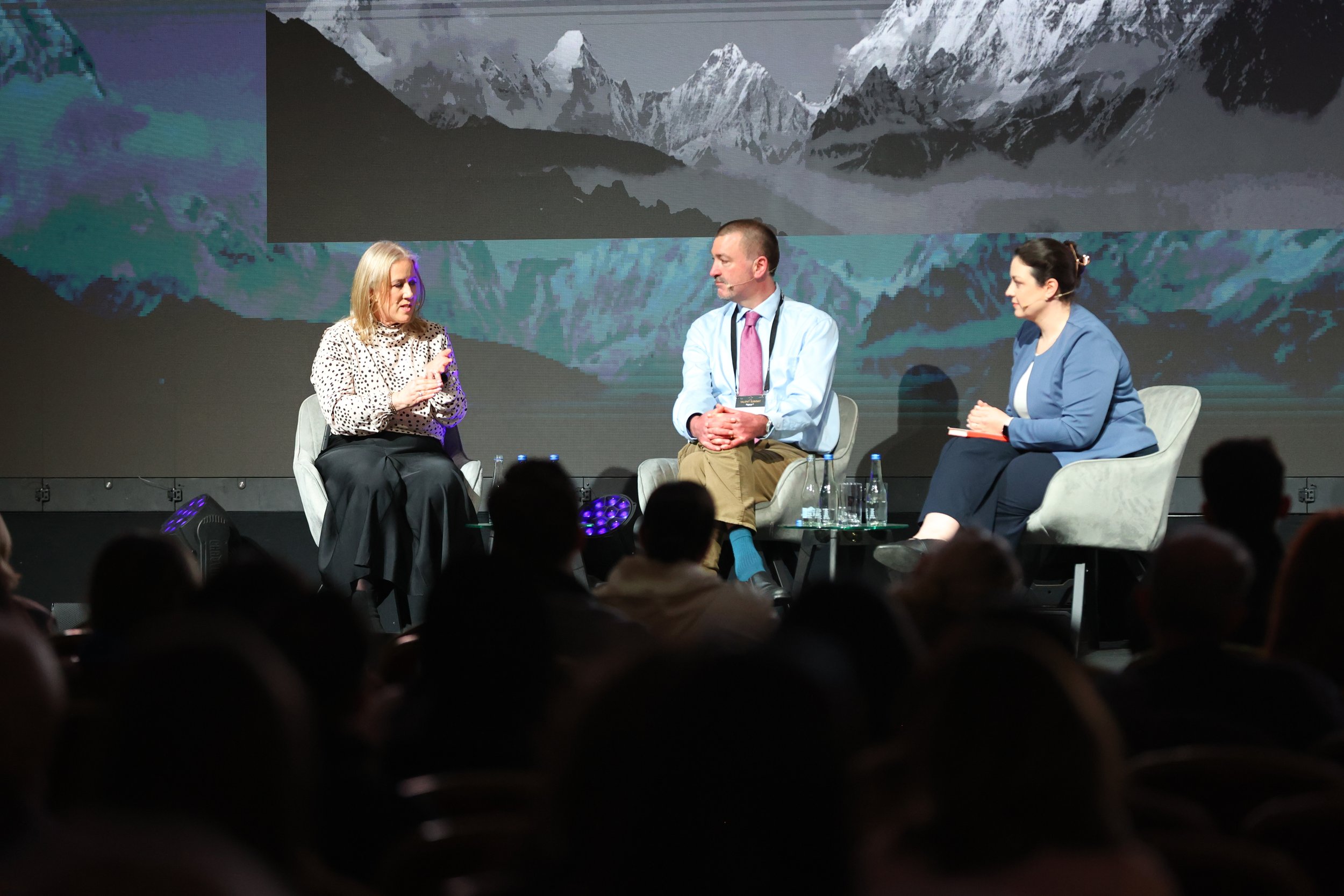 Three people participating in a panel discussion on stage with a mountain landscape projection in the background. The woman on the left is speaking, the man in the middle is listening, and the woman on the right is taking notes.