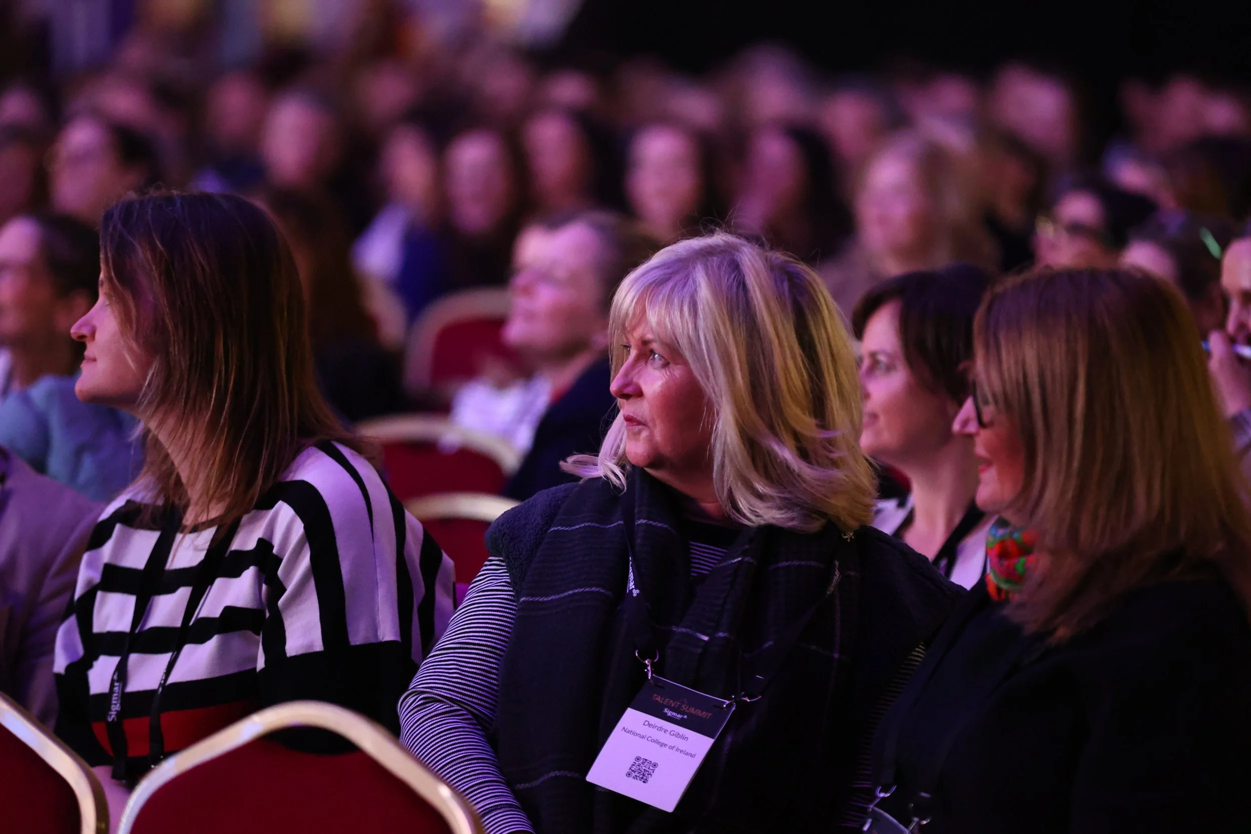 Audience members seated at a conference or event, with focused expressions. The woman in the center has blonde hair, is wearing a striped top and black scarf, with a name badge. Other women are sitting beside her, all paying attention to the proceedi