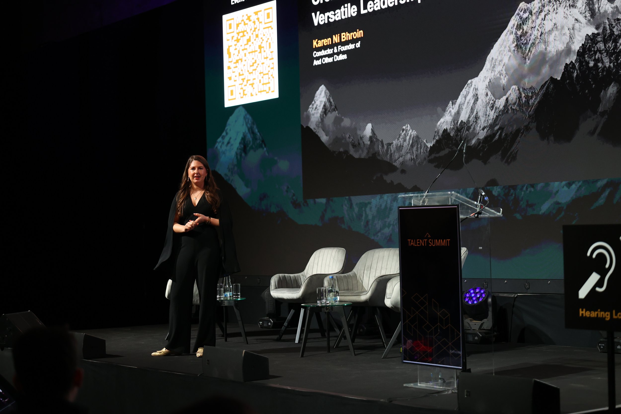 A woman on stage at a conference, standing in front of a large screen displaying mountain images and a QR code. There are three empty chairs with small tables and glasses of water, and a podium with the label 'Talent Summit.' The woman is wearing bla
