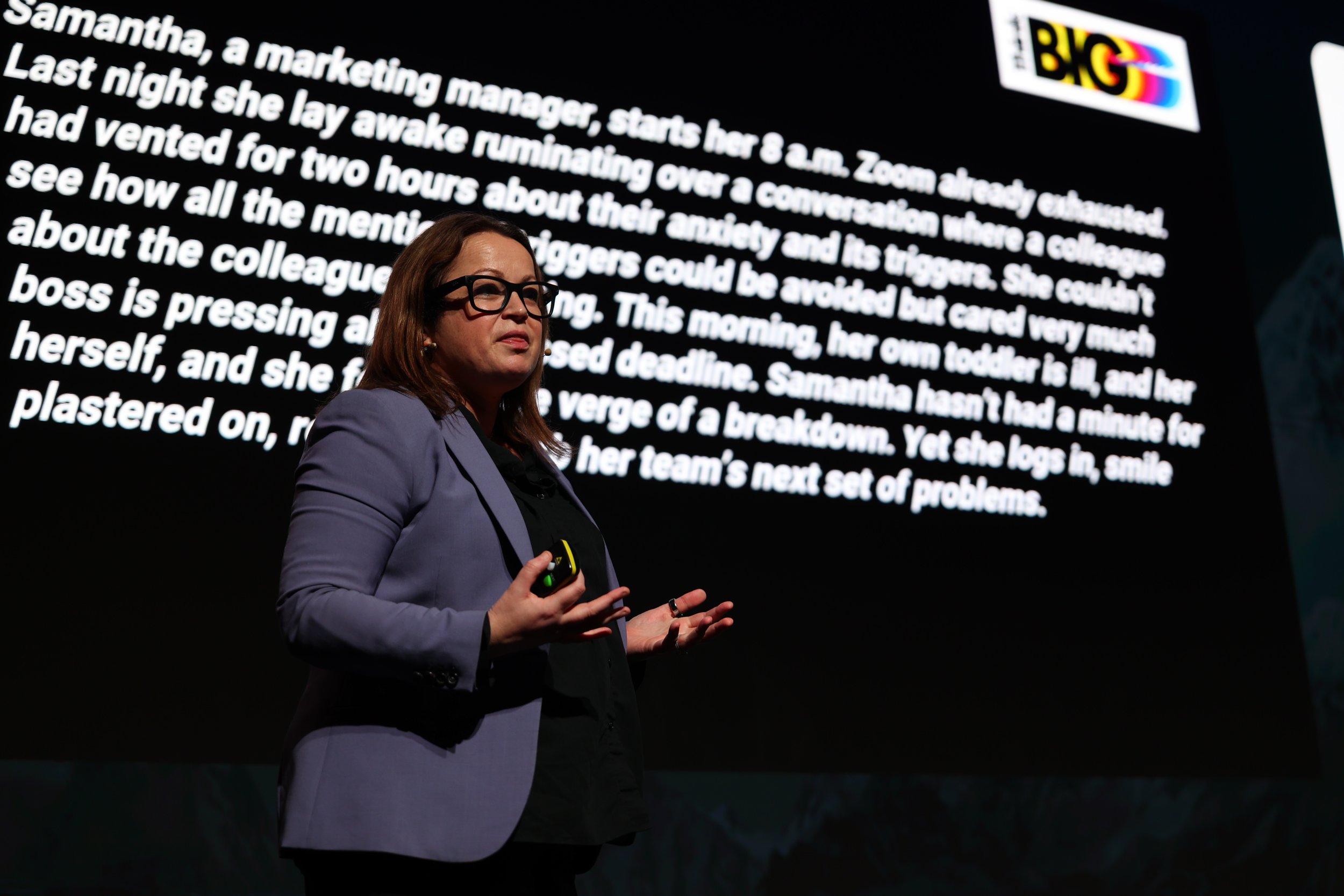 A woman in glasses and a purple blazer giving a presentation in front of a large screen displaying a text paragraph about a person named Samantha. The screen has a logo in the top right corner.