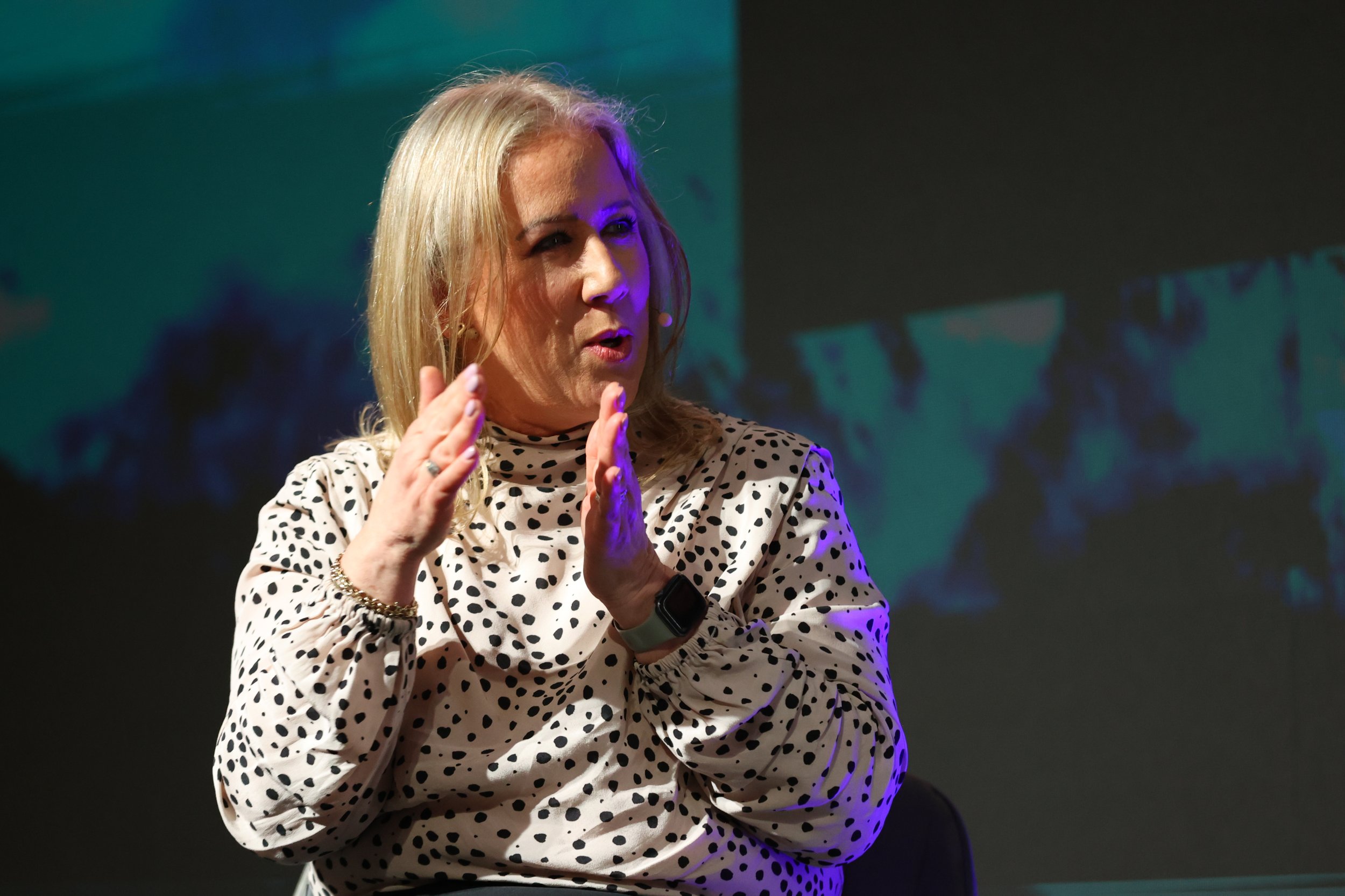 A woman with blonde hair wearing a white blouse with black polka dots, speaking or explaining with her hands raised, seated on a stage with a dark background.
