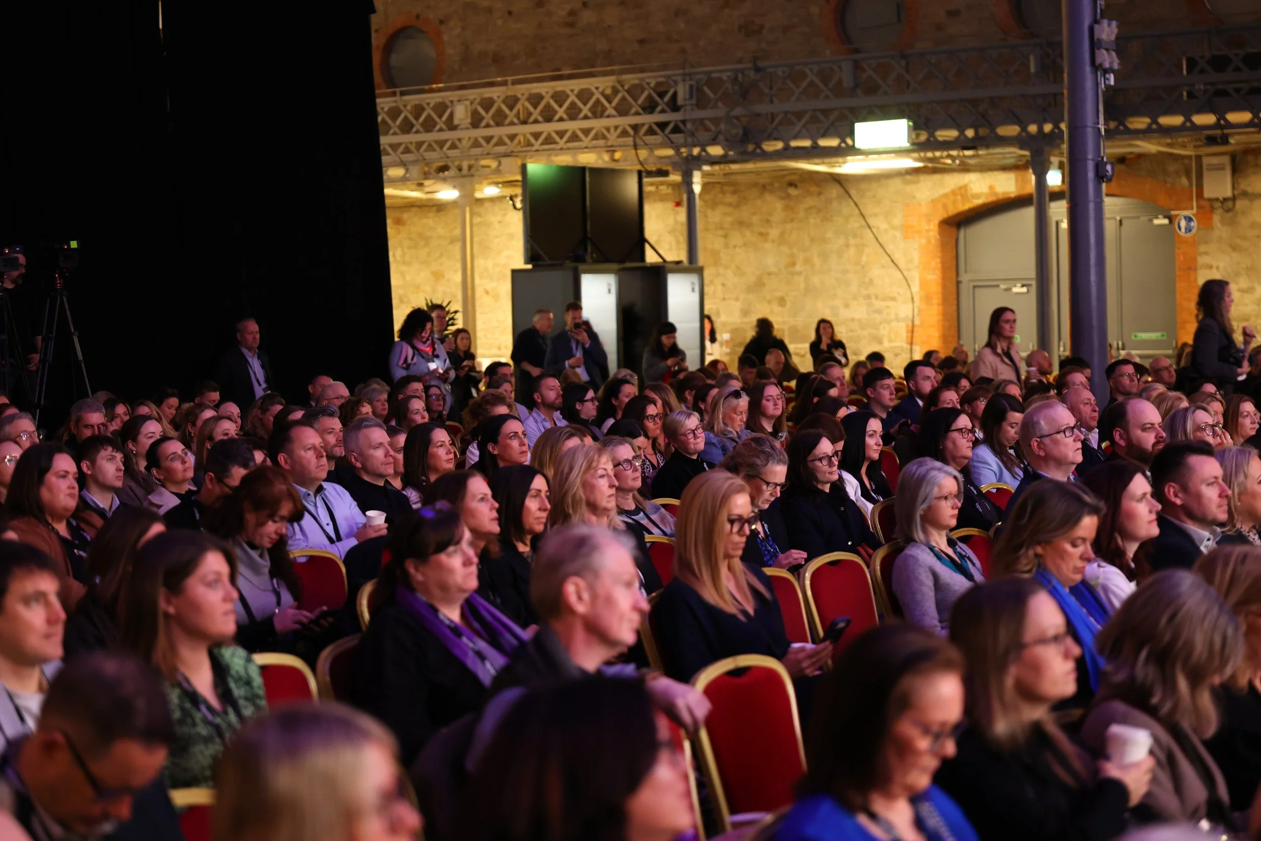 Audience seated in rows at an indoor conference or event, attentively listening to a presentation, with some individuals using smartphones and cups.