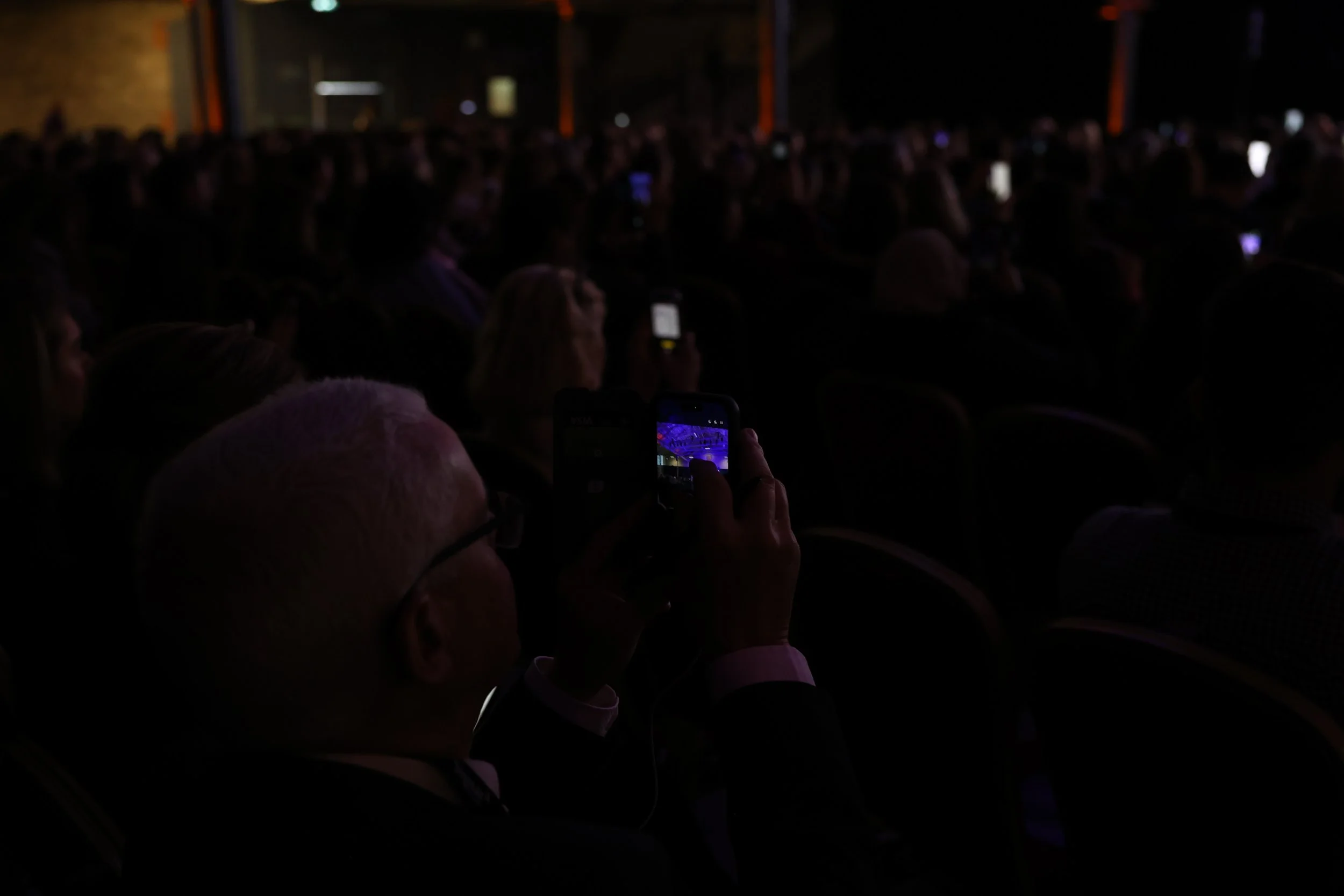 Audience sitting in dark theater, several people using their phones, some screens visible.