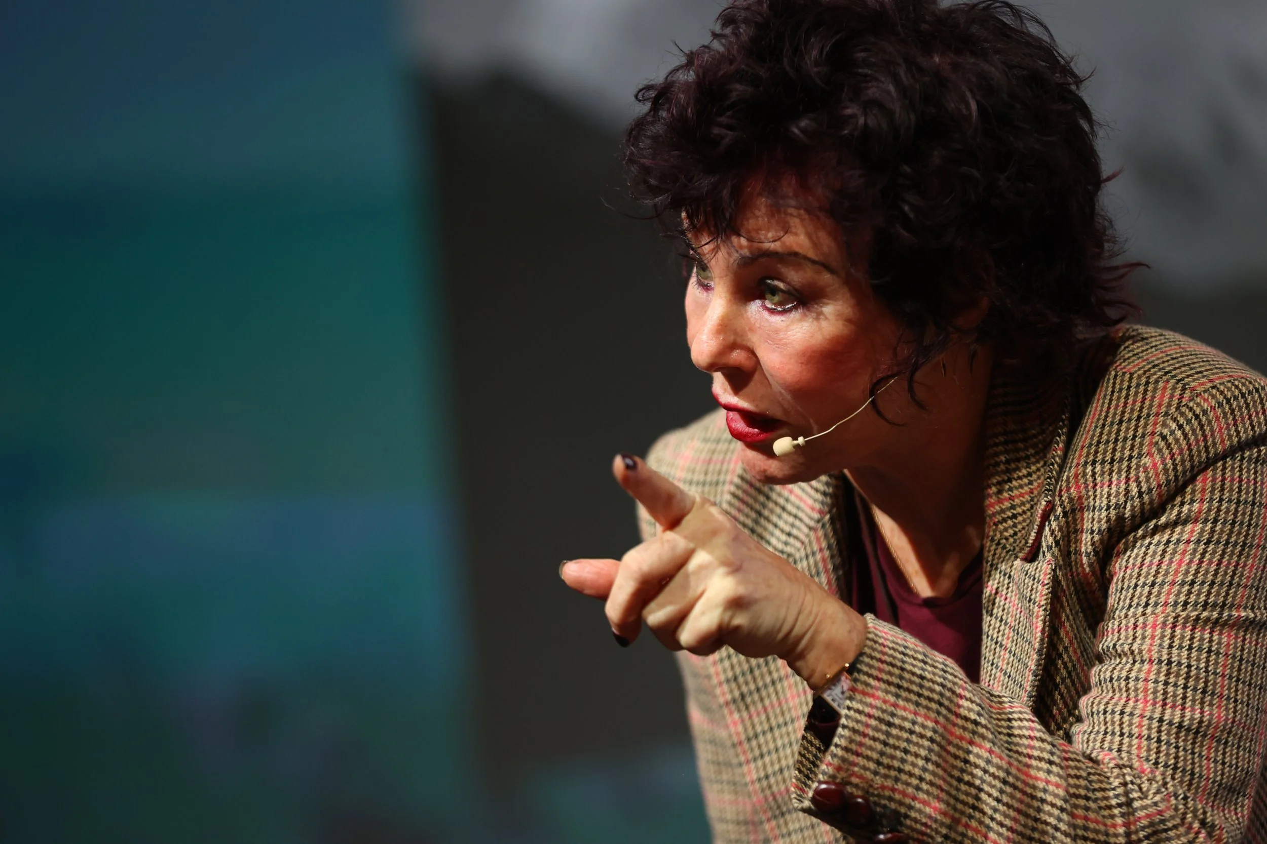 A woman with curly dark hair speaking into a headset microphone, wearing a plaid blazer.