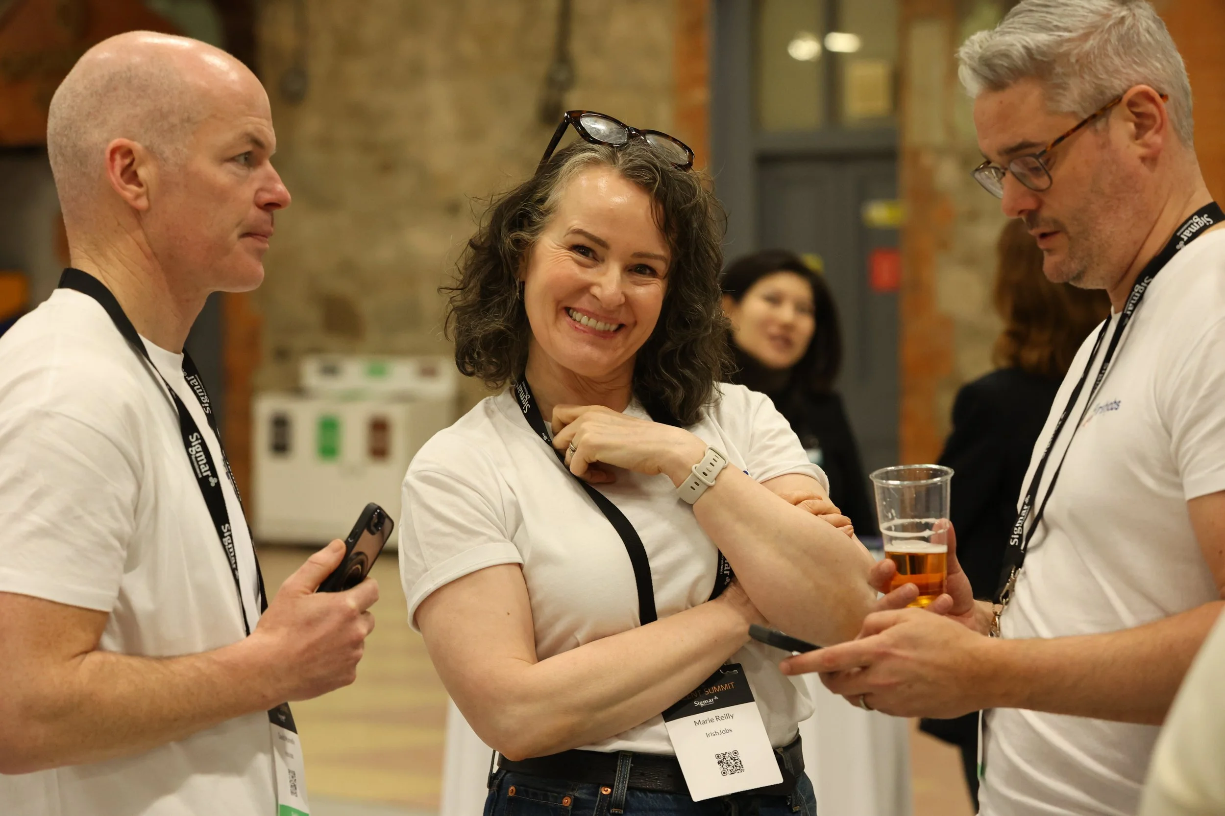 A group of people at a networking event, with a woman smiling and holding a drink, engaged in conversation with two men on either side, all wearing conference badges.