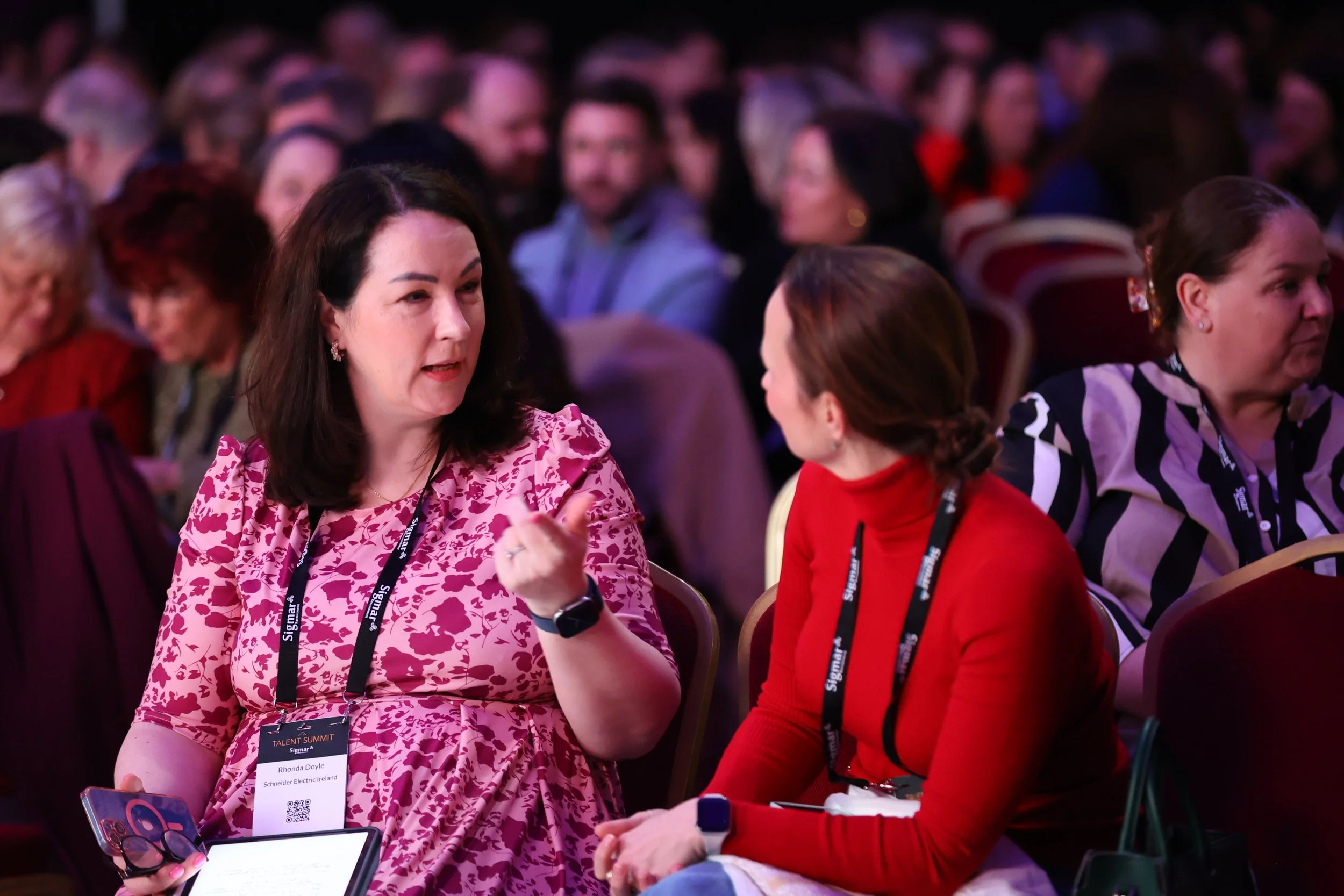 Two women sitting and talking at a conference, with many attendees in the background.