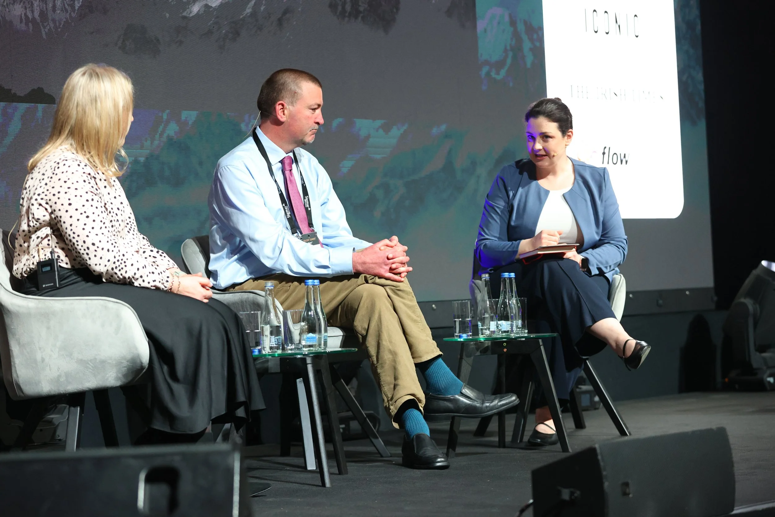 Three people sitting on chairs on a stage during a panel discussion. The woman on the left has blonde hair and is wearing a polka-dot blouse. The man in the middle has short hair, wears a blue shirt with a pink tie, and khaki pants. The woman on the 
