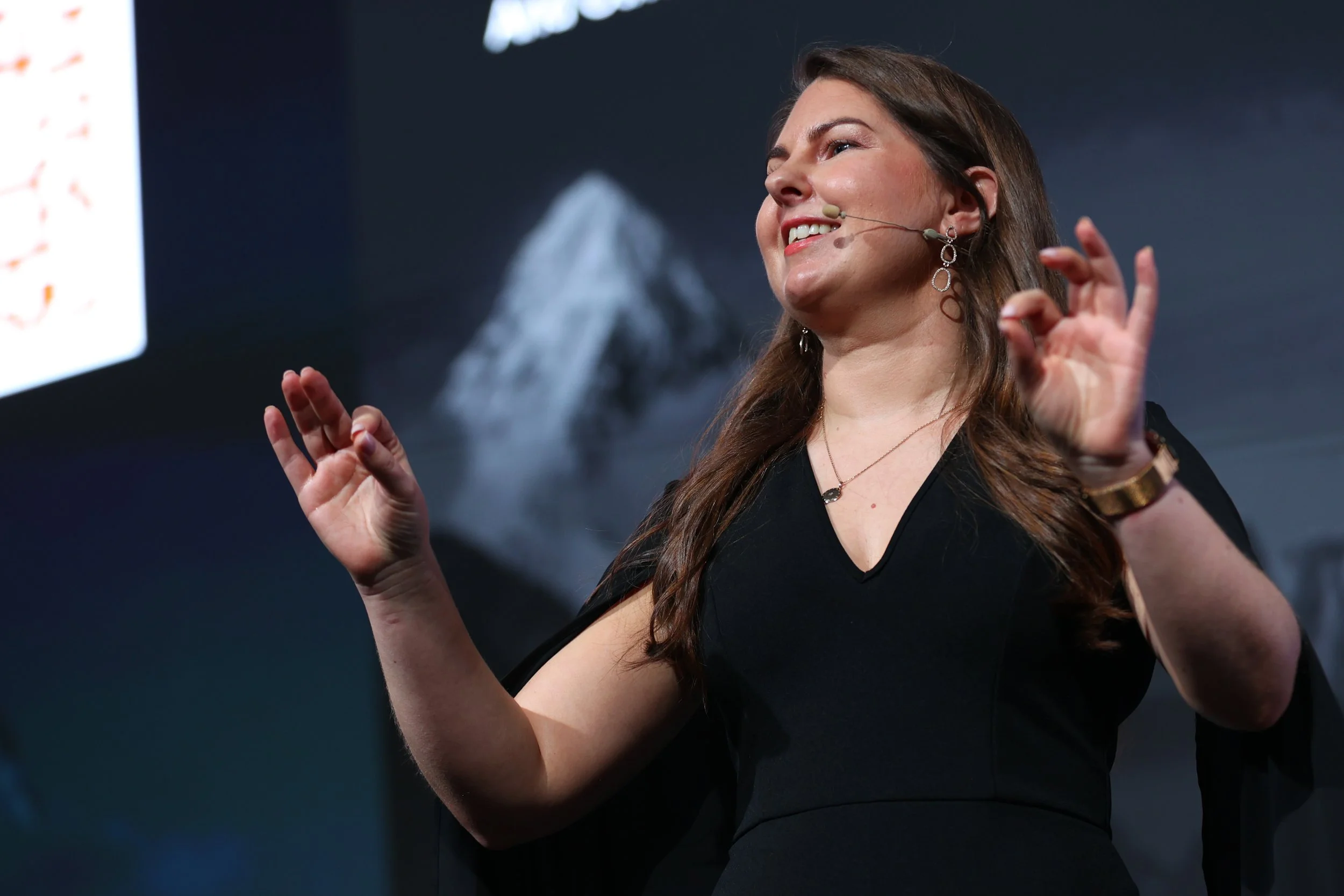 A woman in a black dress giving a presentation, wearing a microphone headset, with a mountain image in the background.
