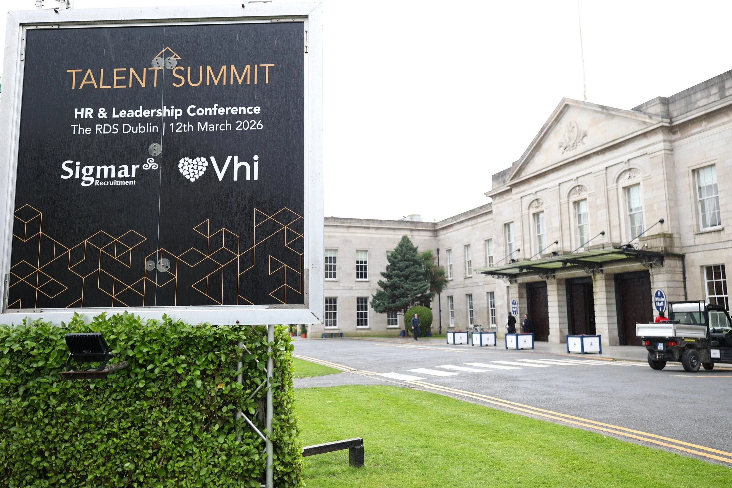 Event sign for Talent Summit HR & Leadership Conference at The RDS Dublin on March 12, 2026, with logos of Sigmar Recruitment and Vhi, outside a large, historic building with a white facade, trees, and a small parked vehicle.