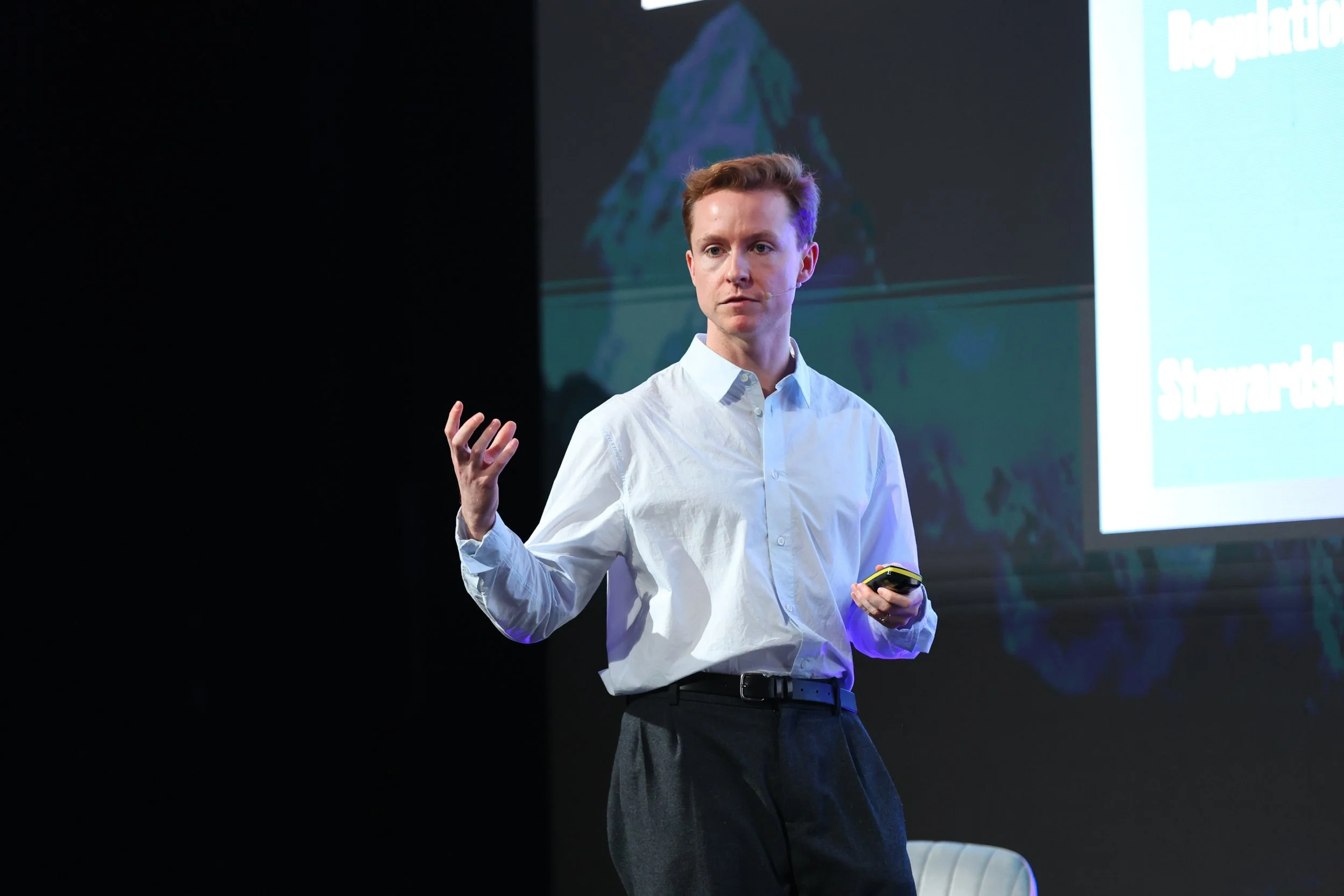Young man with red hair in a white shirt giving a presentation on stage, holding a remote control in his right hand, with a large screen behind him displaying a slide.