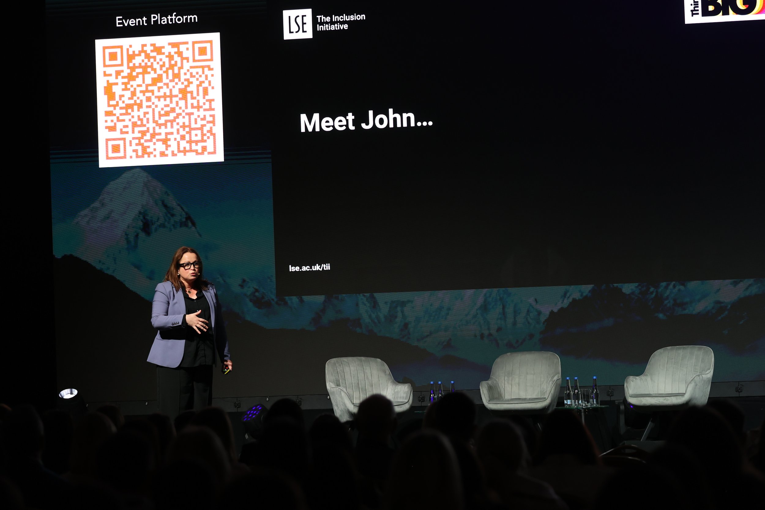 A woman speaking on stage during a conference, with a large screen behind her displaying the text 'Meet John...' and a QR code. The stage has three empty chairs and some bottles of water. The slide features the logos of LSE and The Inclusion Initiati