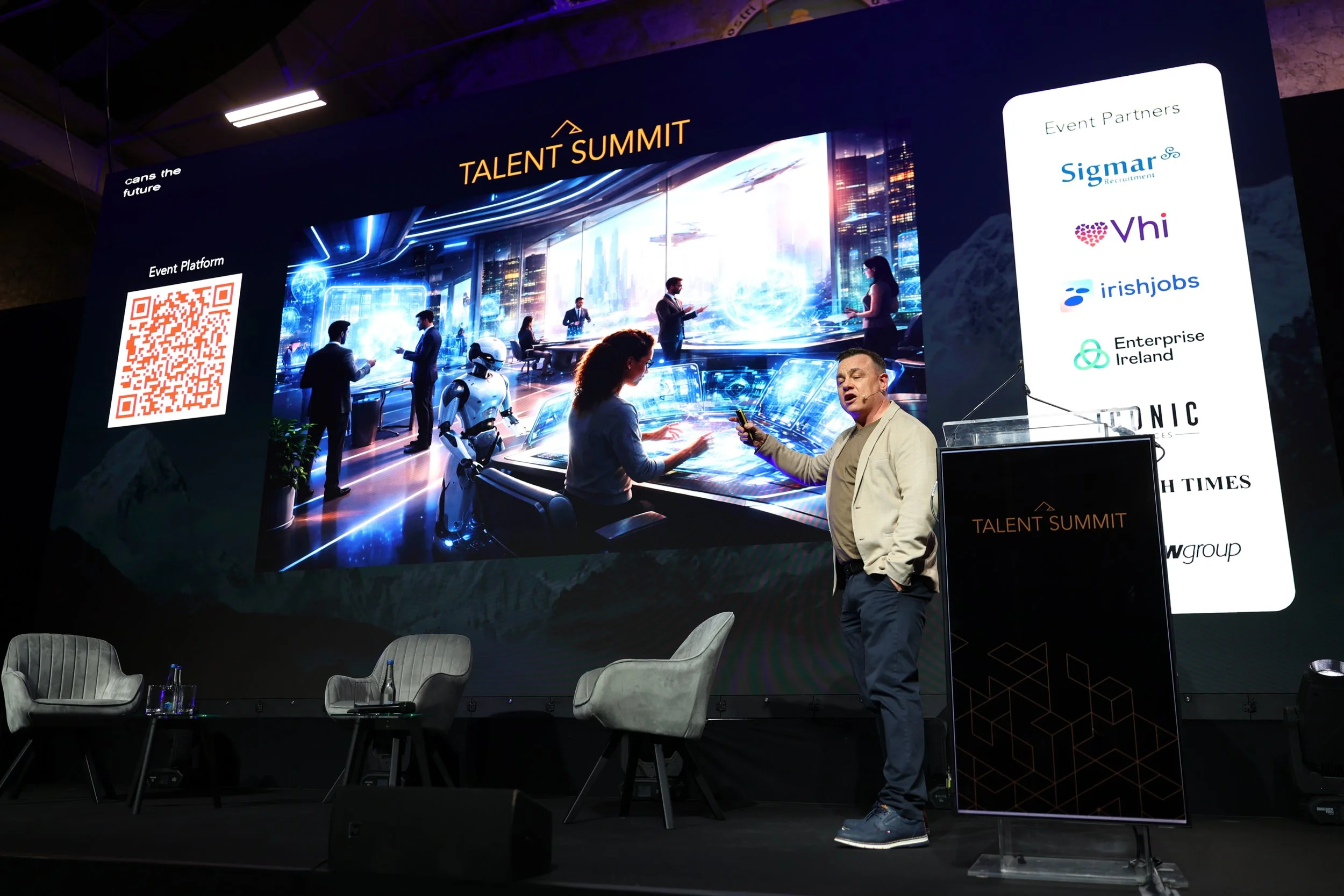 A man on stage at a conference called Talent Summit, speaking and pointing with a laser pointer while a large screen behind him displays a futuristic city scene with people, robots, and technology. There are chairs on stage and a sign with event part