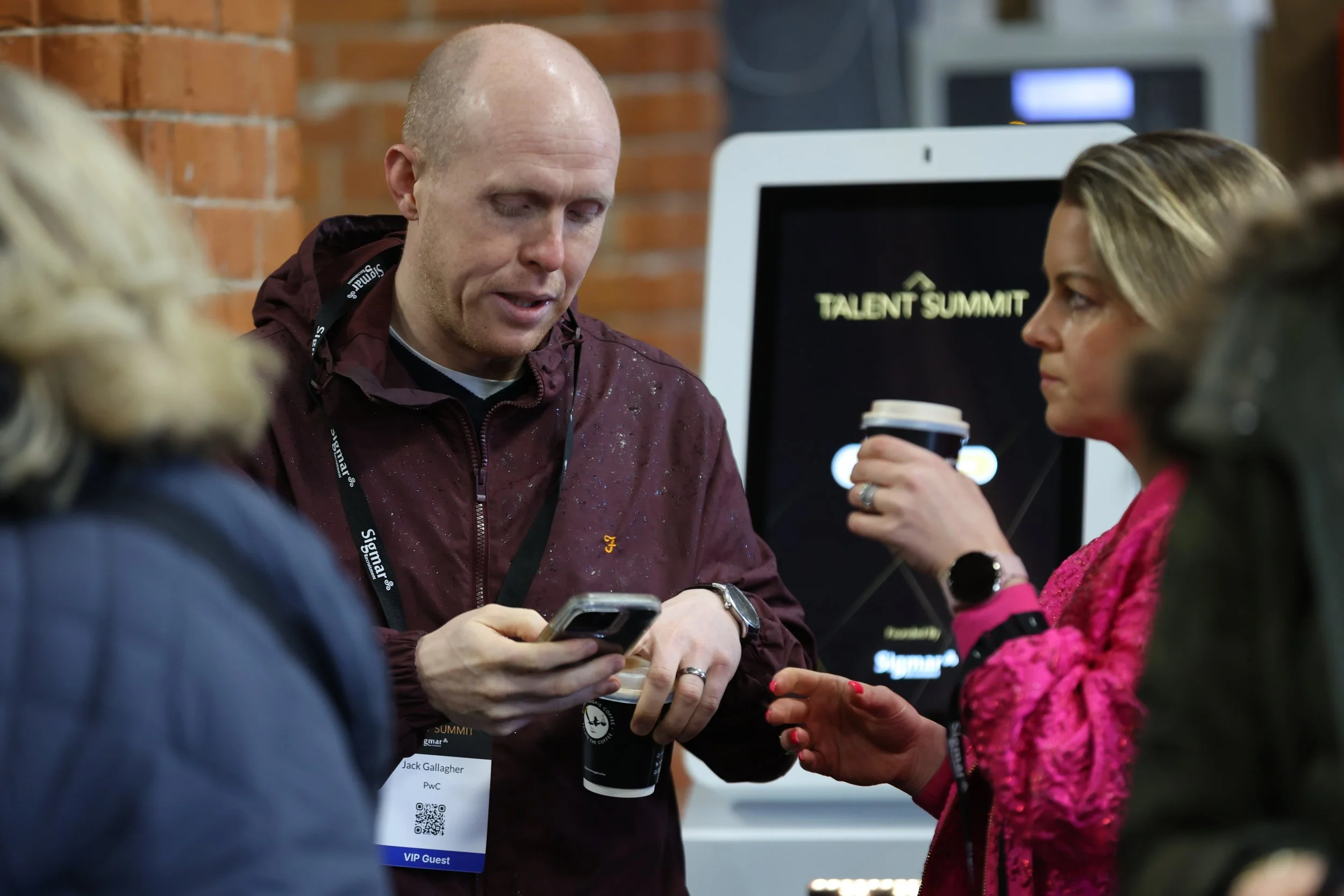 A group of people at an event called 'Talent Summit'. A man with a badge holding a phone and a woman with a cup of coffee engaging in conversation. The background has a screen displaying the event name.