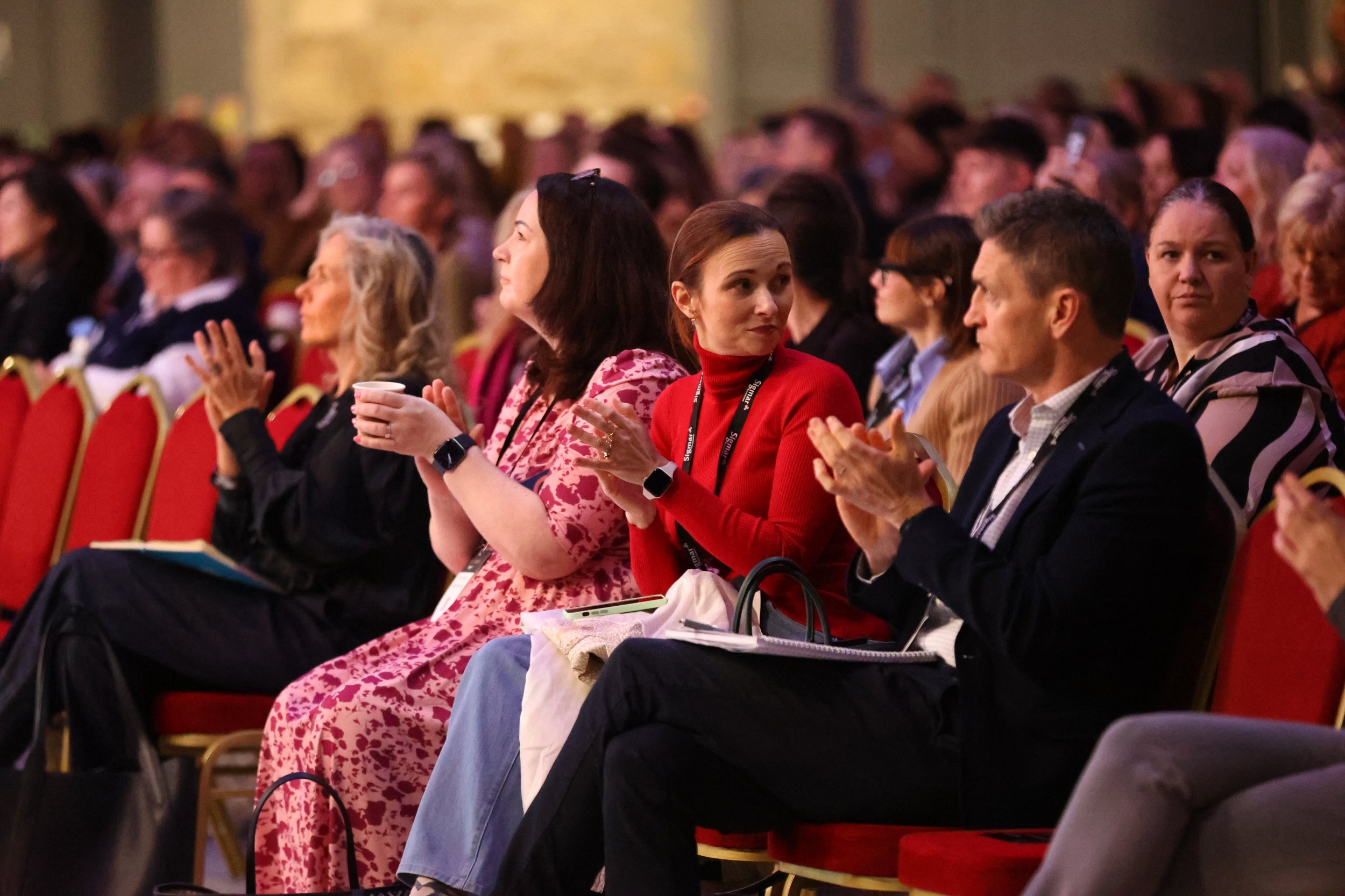 Attendees at a conference seated in rows of red chairs, listening and clapping.