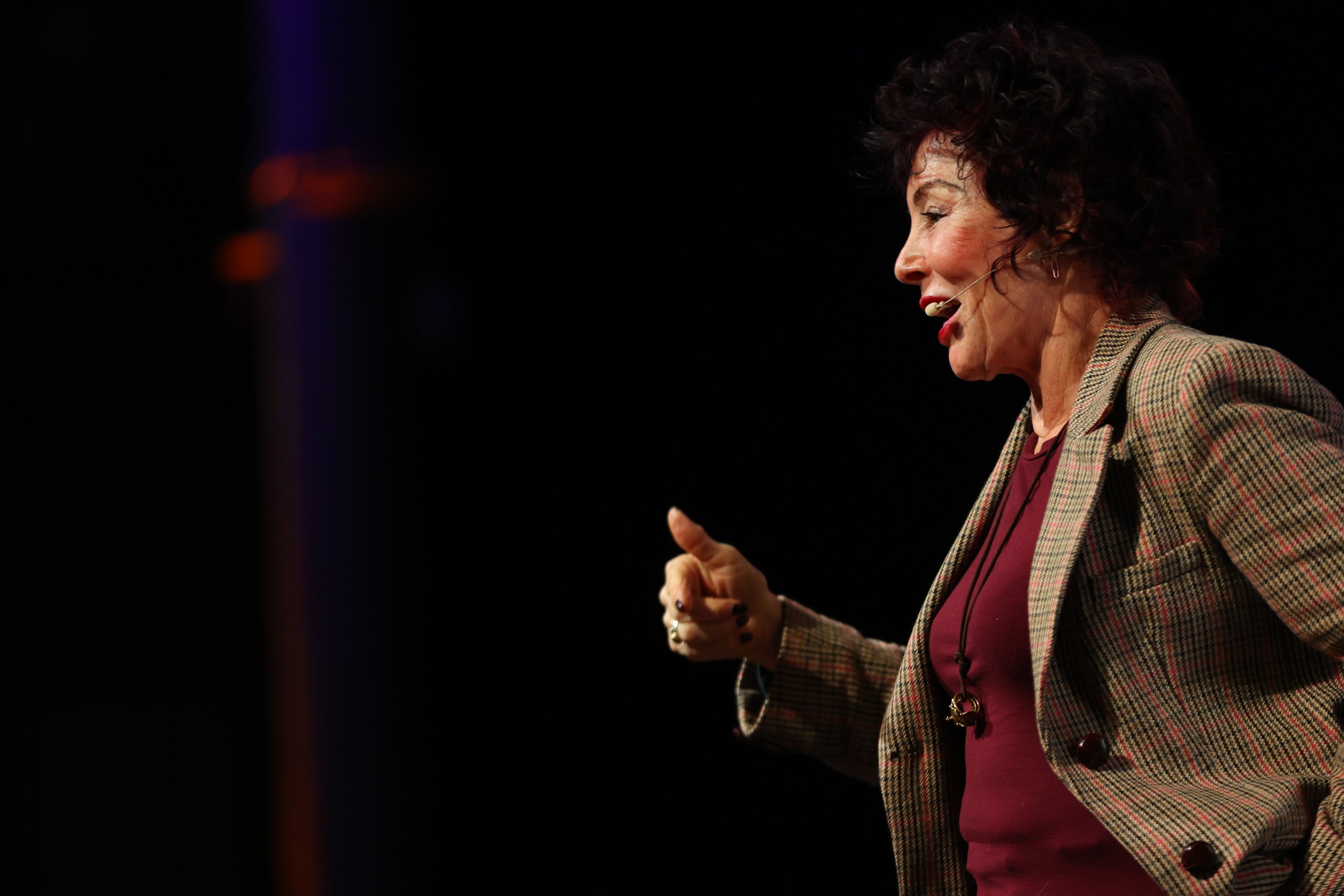 A woman with curly dark hair, wearing a plaid blazer and burgundy top, is speaking on a stage with a dark background.