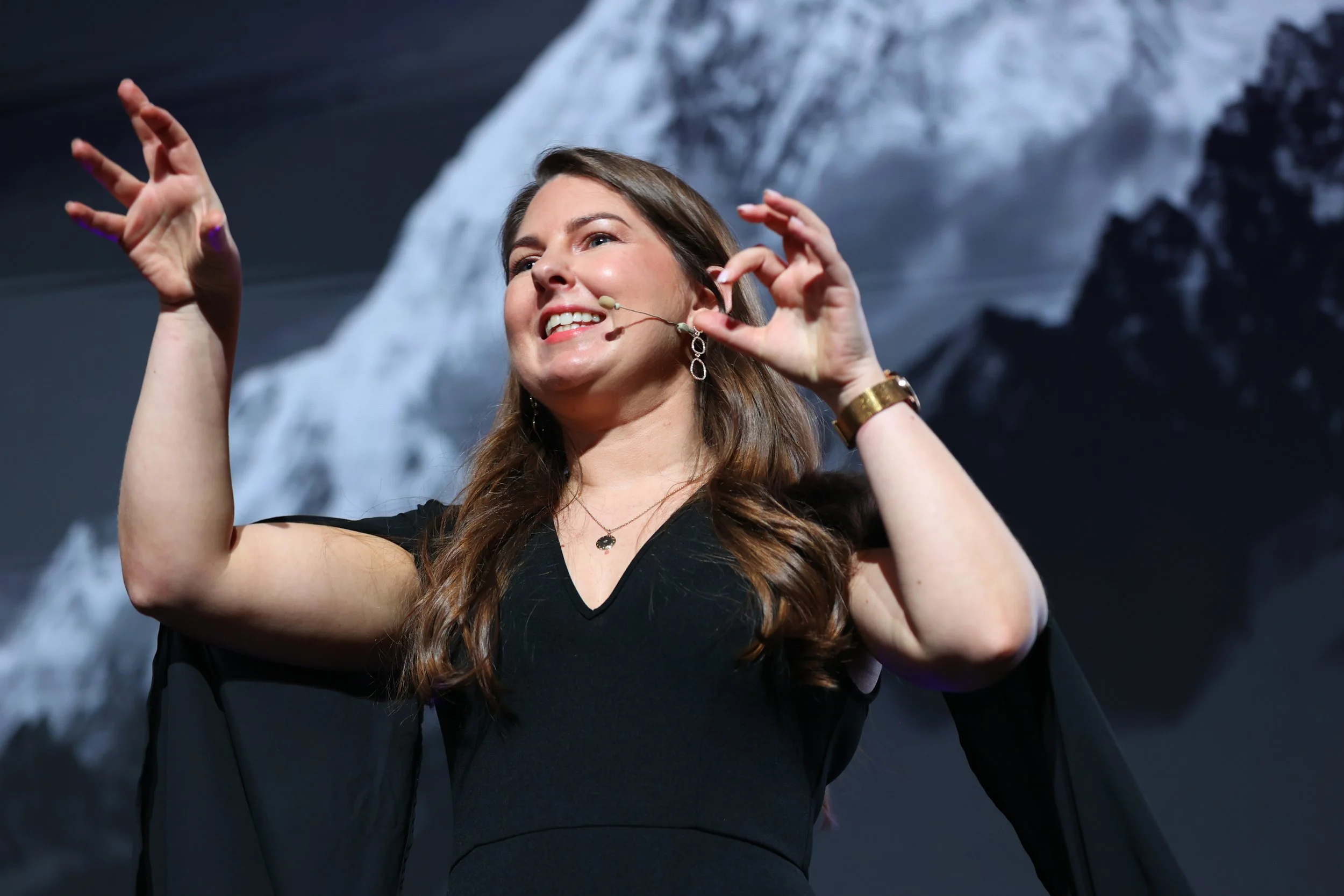 A woman with long brown hair wearing a black dress and earrings, smiling and gesturing with her hands on stage, with a mountain landscape in the background.