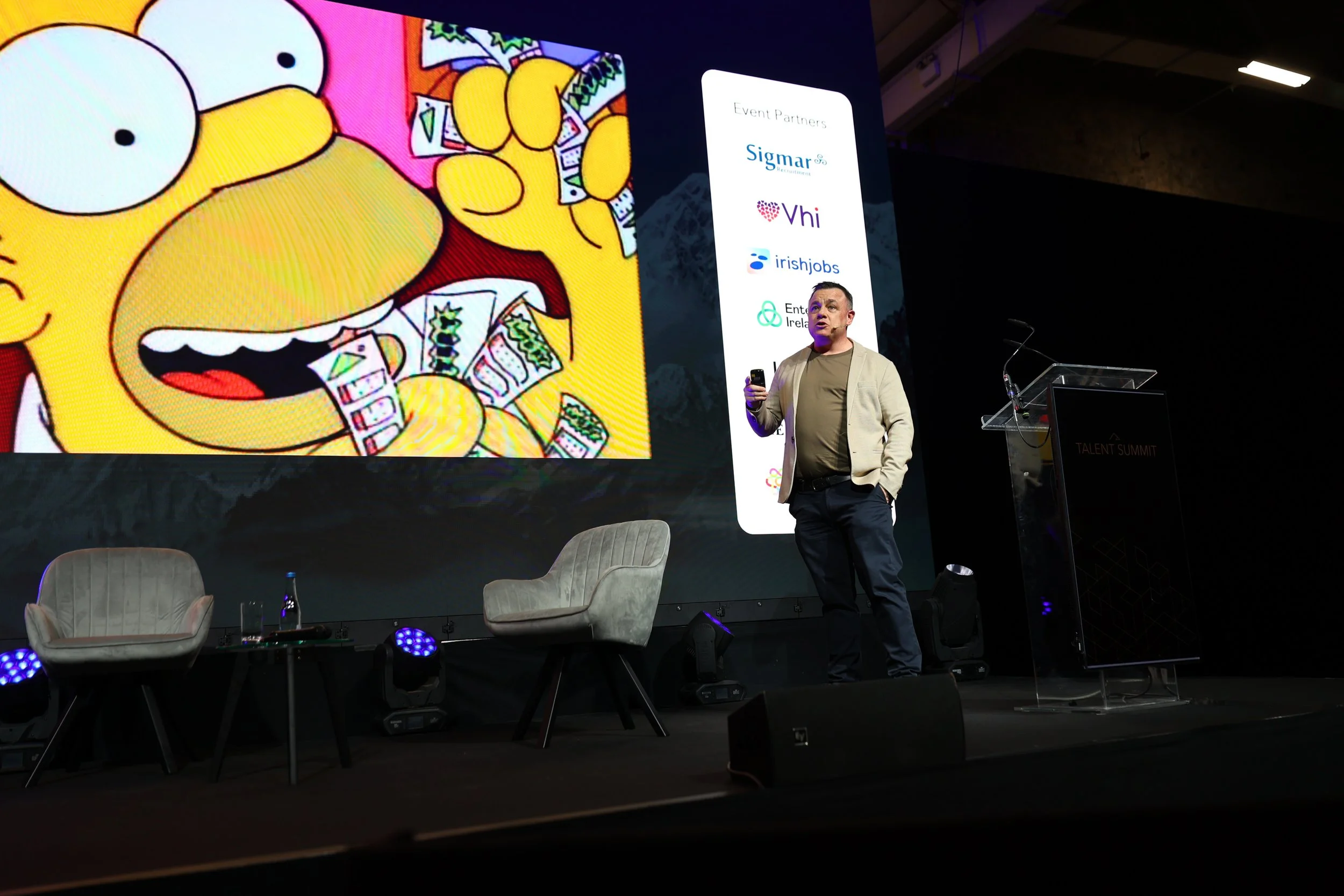 A man giving a presentation on a stage with a large screen behind him displaying a cartoon character of a yellow animal with large eyes and a wide-open mouth, holding and throwing money. The stage has two chairs, a small table with a water bottle, an