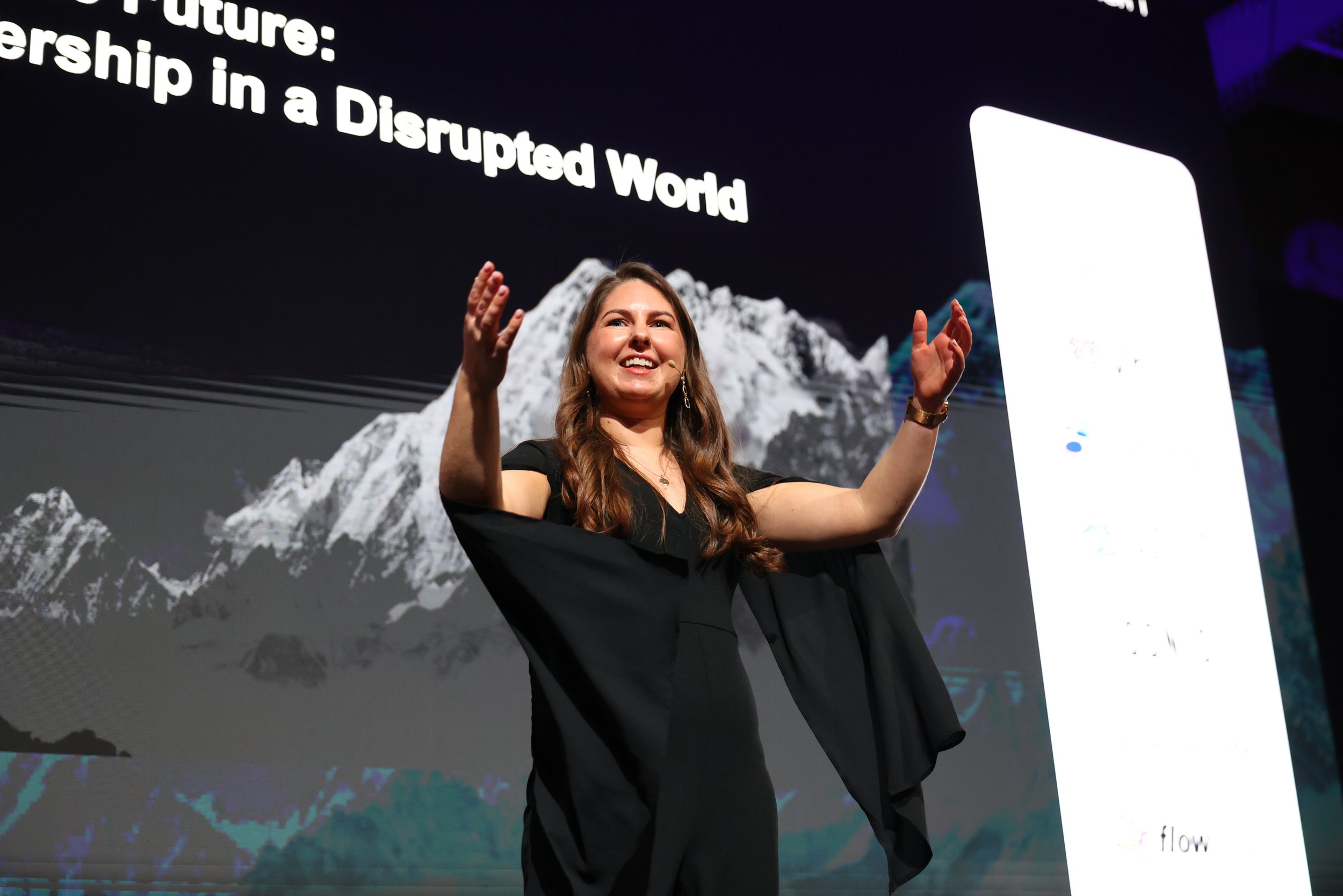 A woman standing on a stage with a large screen behind her. The screen displays a mountain image and text about future leadership in a disrupted world. She is smiling, wearing a black dress, and gesturing with both hands.