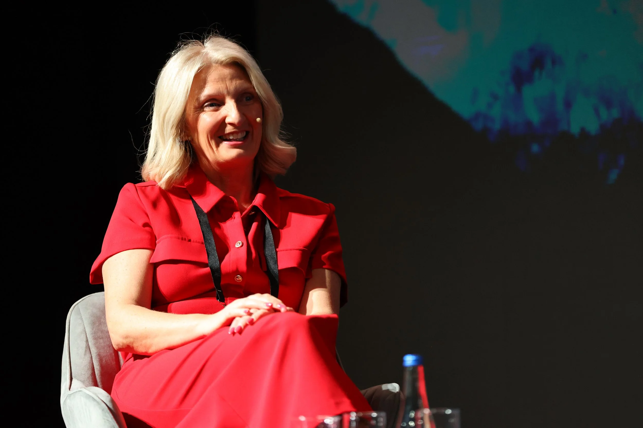 Woman with blonde hair wearing a red dress, sitting on a stage with a dark background and a small part of a screen visible behind her.