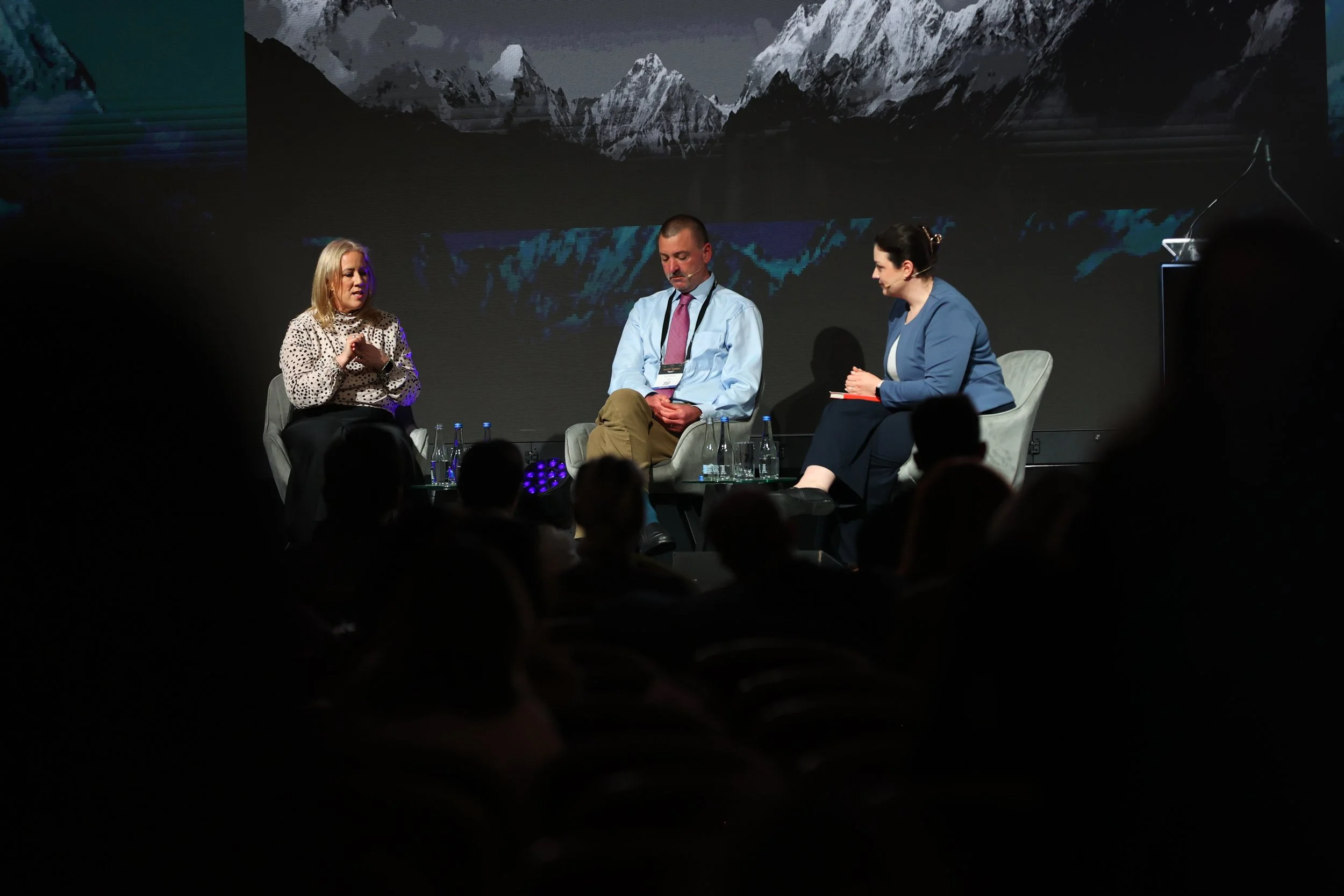 A panel discussion with three people on stage, two women and one man, seated in chairs, with an audience in front of them. The background shows a mountainous landscape.