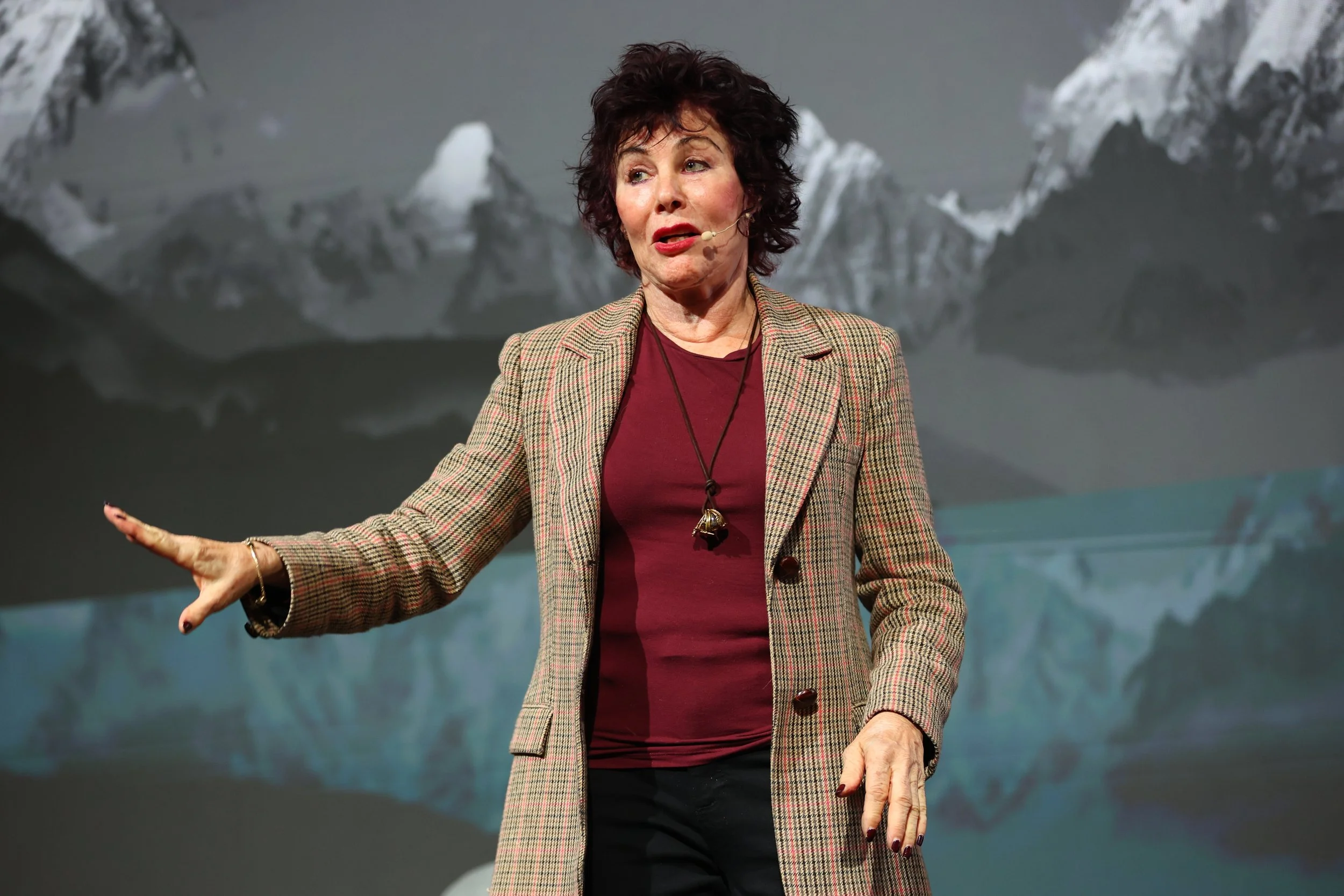 A woman with short curly hair, wearing a checkered blazer and a microphone headset, speaking on stage with a mountain landscape background.