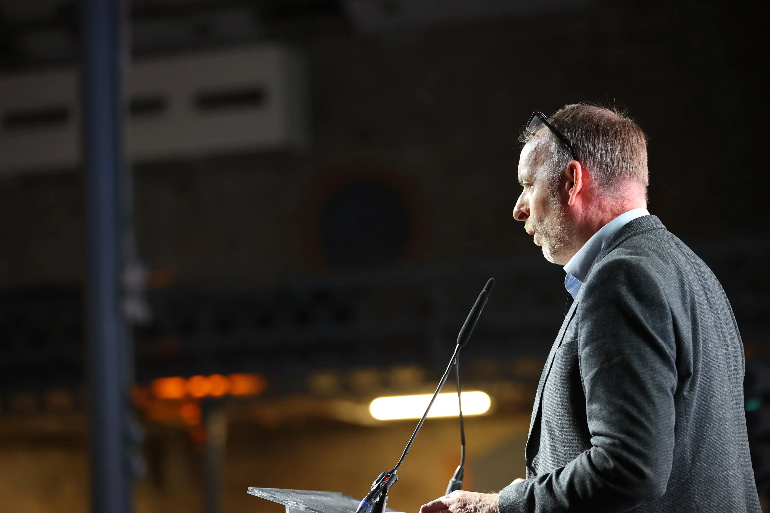 A middle-aged man with a beard and glasses resting on his head, dressed in a gray blazer and white shirt, speaking at a podium with microphones, indoors with dim lighting.