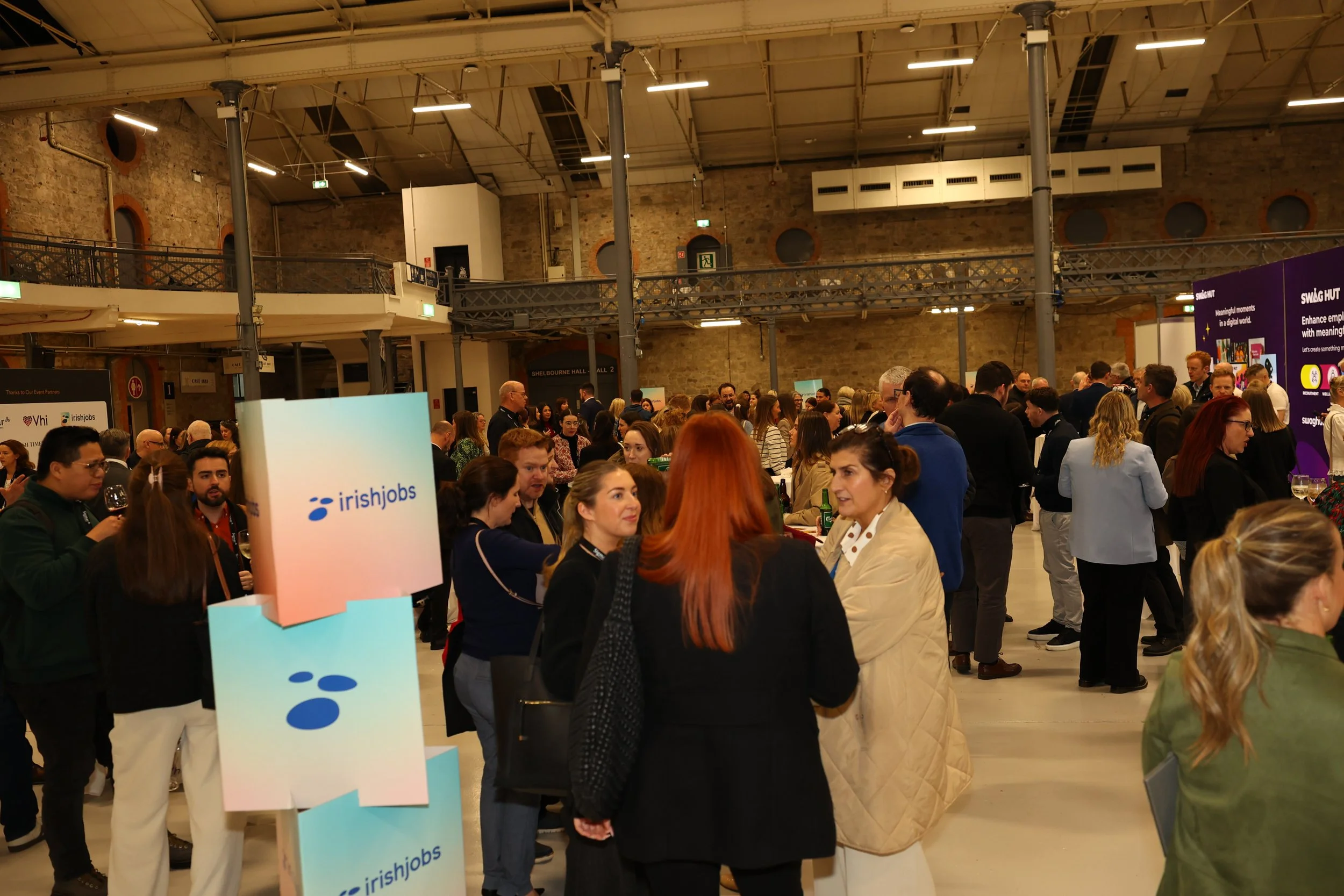 A large indoor event hall filled with a diverse crowd of people attending a professional networking or conference event. There are various booths and signs, including one that reads IrishJobs. The hall has exposed brick walls, high ceilings, and brig
