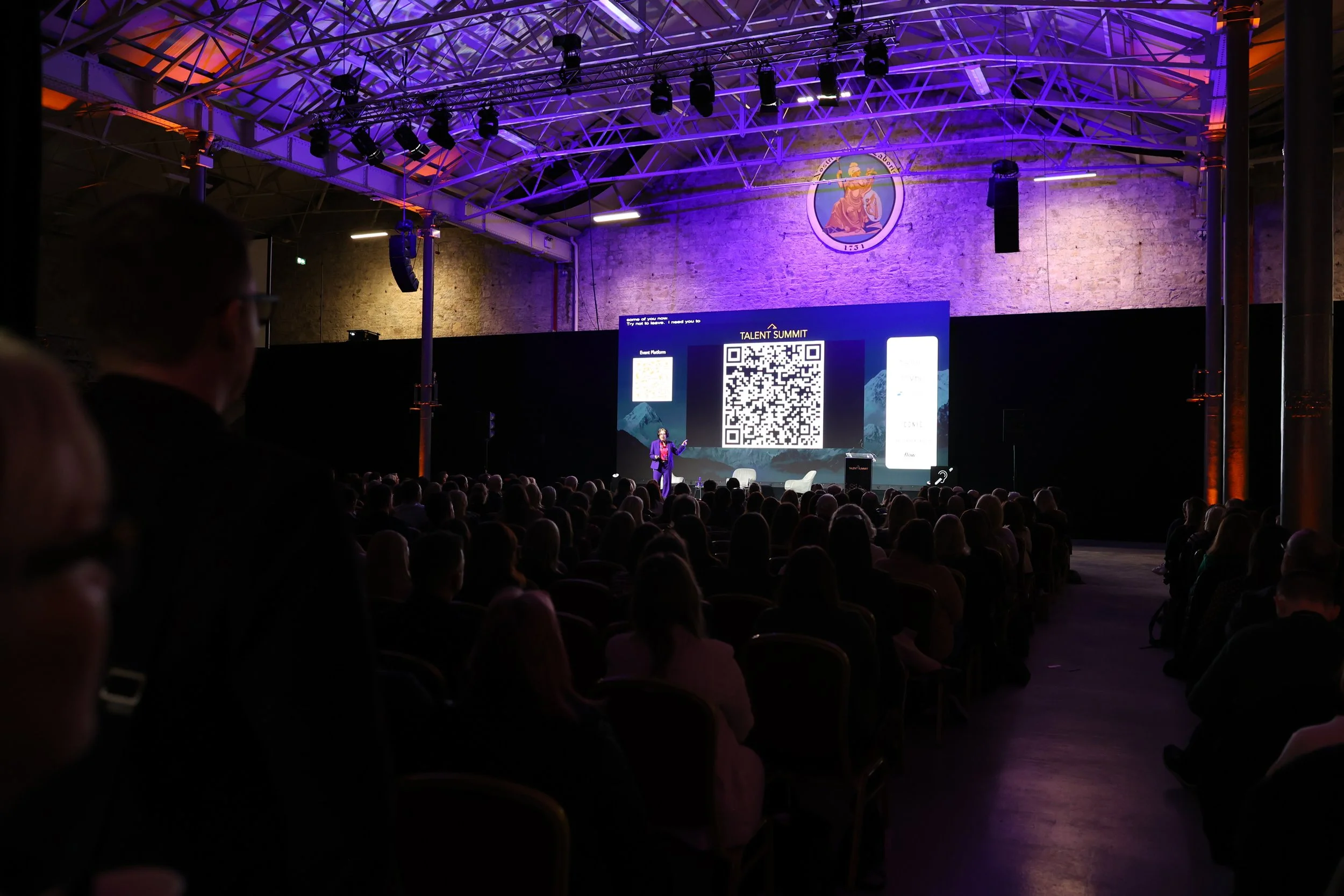 A conference or seminar taking place in a large, industrial-style venue with a high ceiling and exposed brick walls. A presenter stands on stage in front of a large screen displaying a QR code and the words 'Talent Summit.' An audience of seated atte