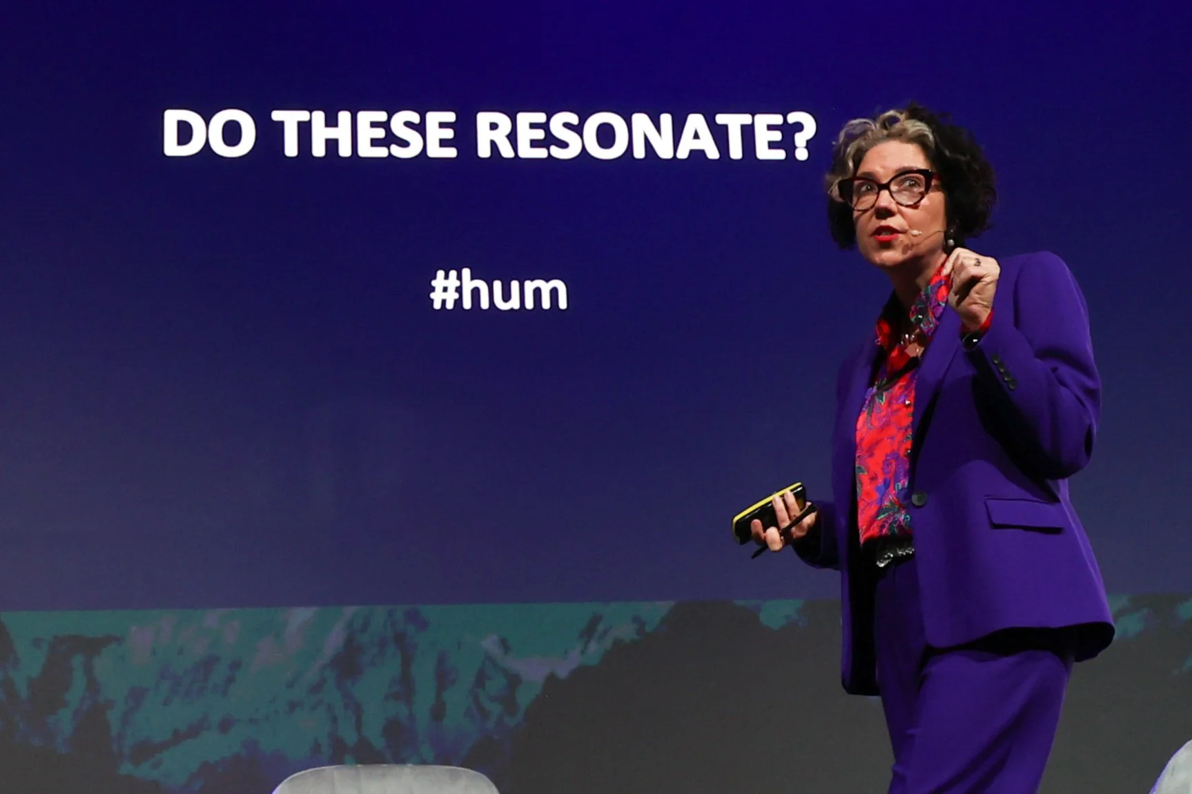 A woman in a purple suit and glasses speaking on stage with a dark background, showing text that asks 'Do these resonate?' and the hashtag '#hum'.