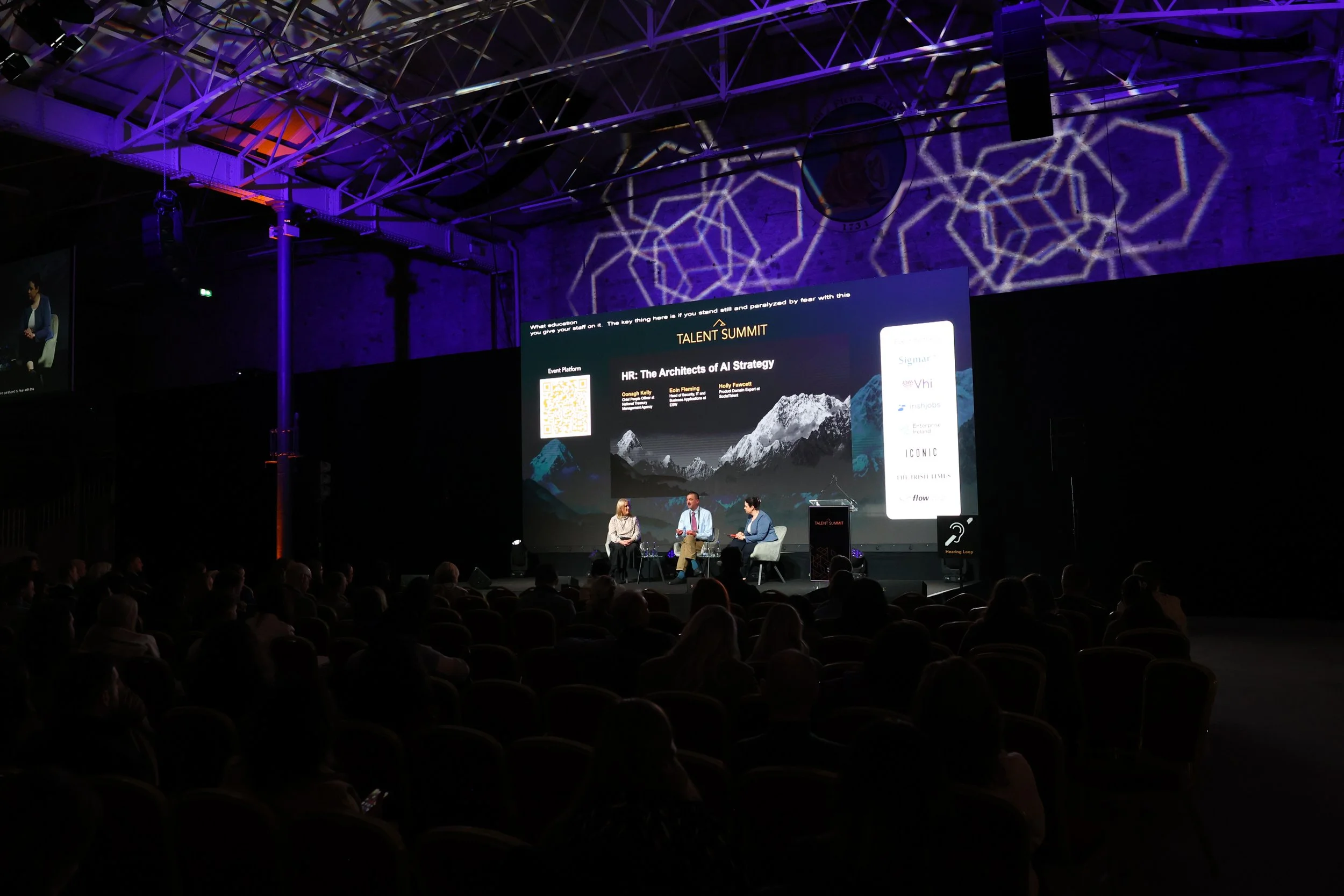 An audience watching a panel discussion at a conference with a large digital screen displaying the topic 'HR: The Architects of AI Strategy' at the Talent Summit, inside a dark auditorium with purple lighting and geometric light projections on the ce